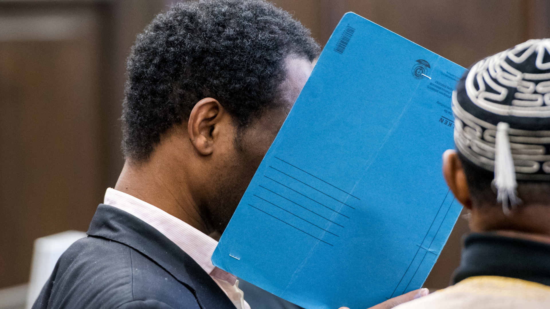 Der wegen zweifachen Mordes angeklagte 34-Jährige steht im Landgericht und hält sich eine Mappe vor sein Gesicht. | Bildquelle: dpa