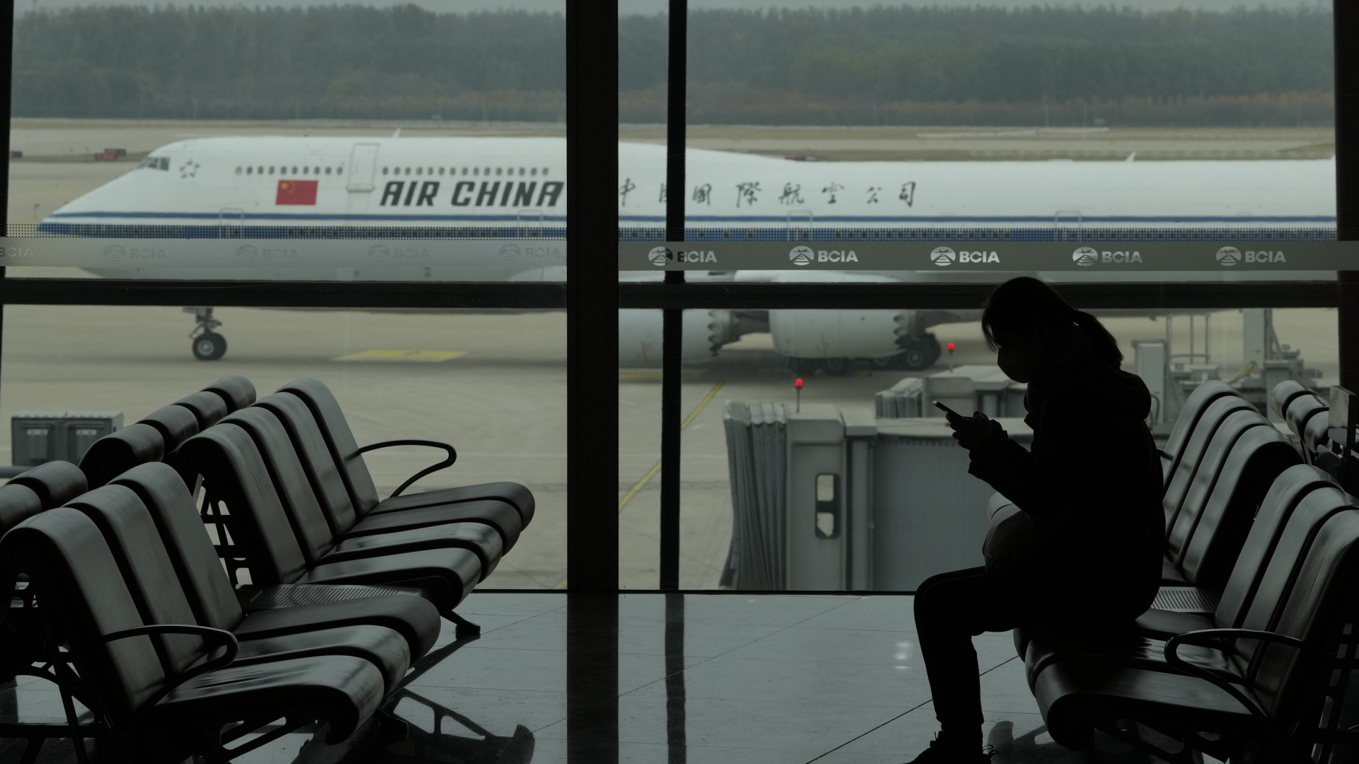 Eine Frau sitzt am Flughafen am Gate, im Hintergrund ist ein chinesisches Flugzeug zu sehen. | AP Photo/Ng Han Guan