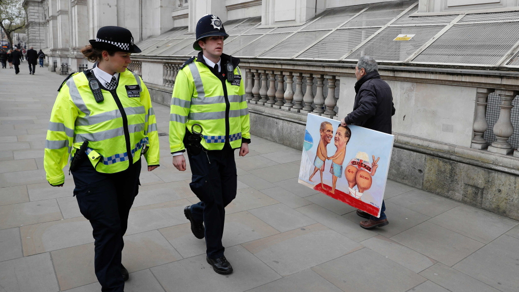 Zwei Londoner Polizisten gehen an einem Mann mit einem Gemälde vorbei. Darauf eine Karrikatur von Us-Präsident Obama und Englands Premier Cameron.