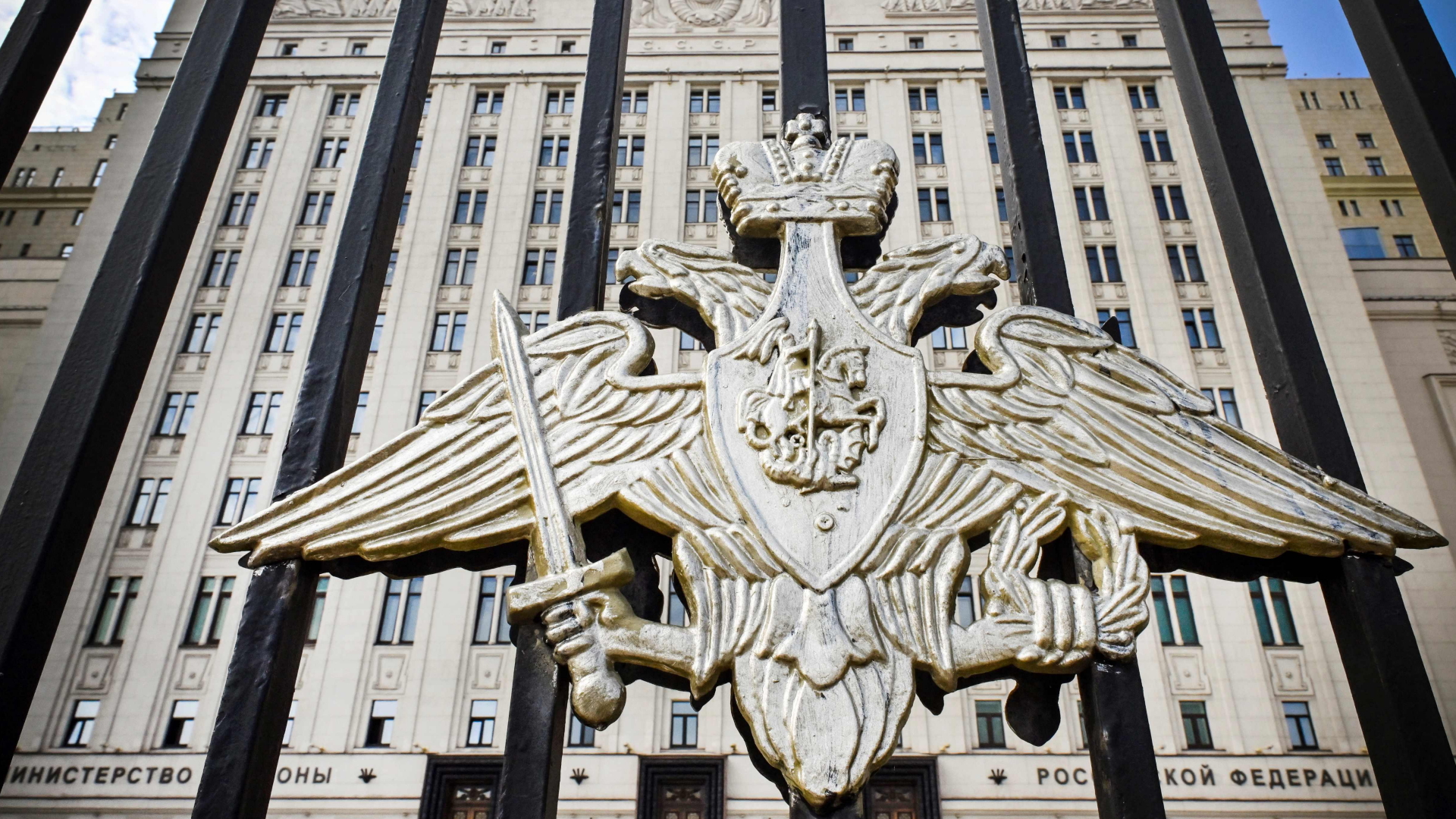 Golden anmutendes Wappen auf einem schwarzen Tor, im Hintergrund ein sehr großes Gebäude: Das russische Verteidigungsministerium. | AFP