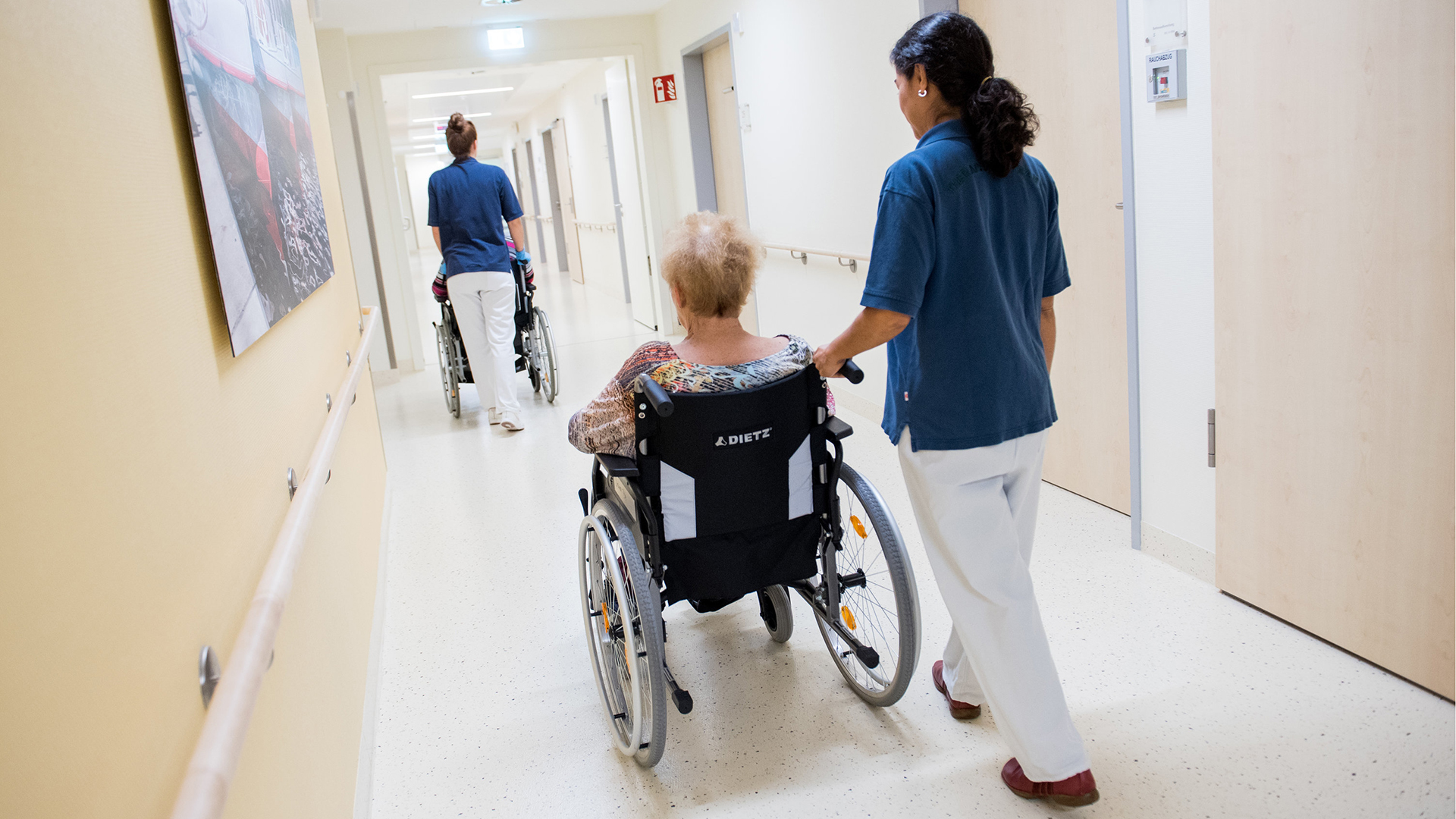 Zwei Pflegerinnen schieben Patienten mit Rollstühlen durch einen Flur.