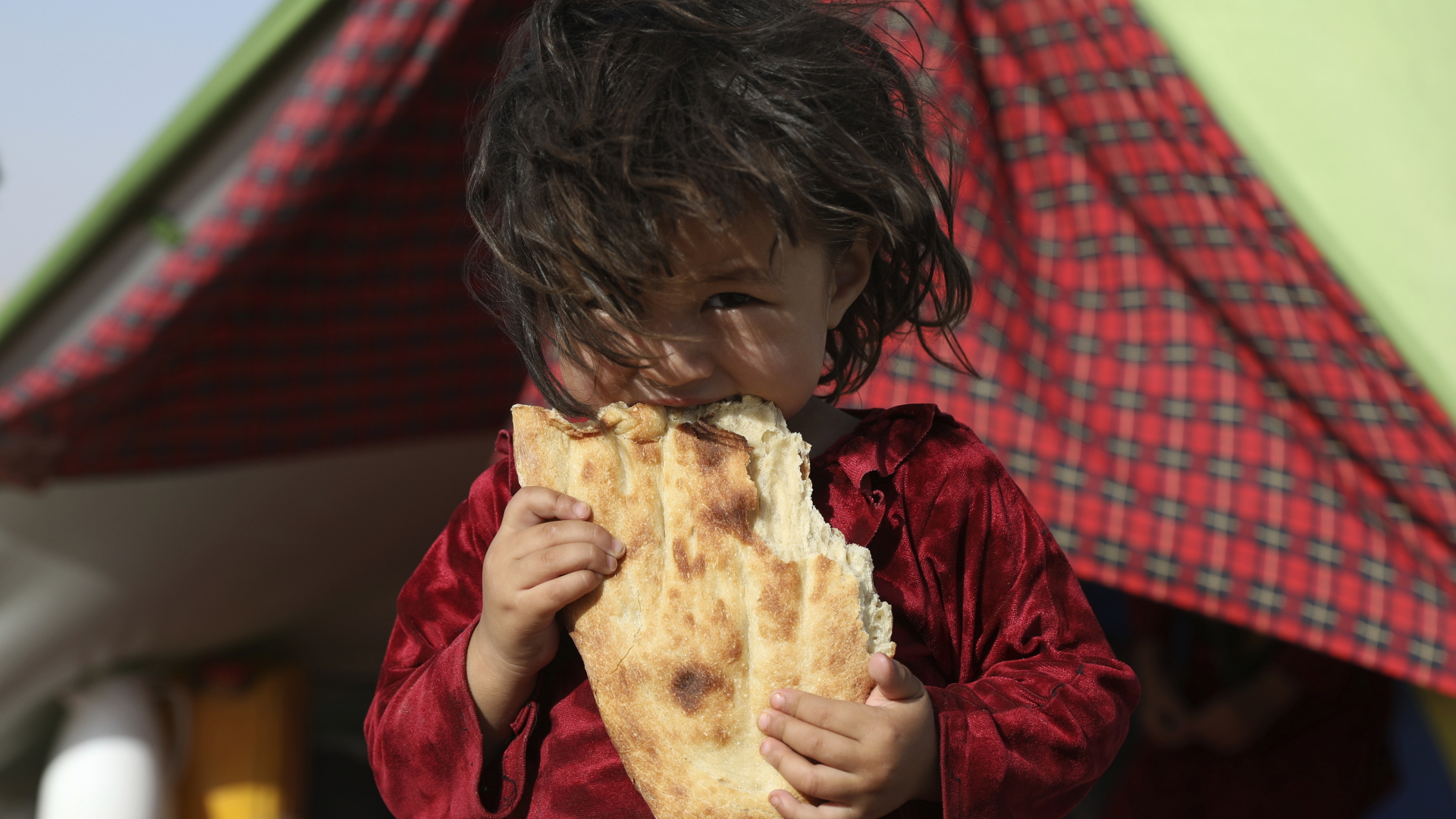 Ein afghanisches Mädchen, das aufgrund von von Kämpfen zwischen den Taliban und afghanischen Sicherheitskräften aus ihrer Heimat geflohen ist, steht vor einem Zelt und isst Brot. | dpa