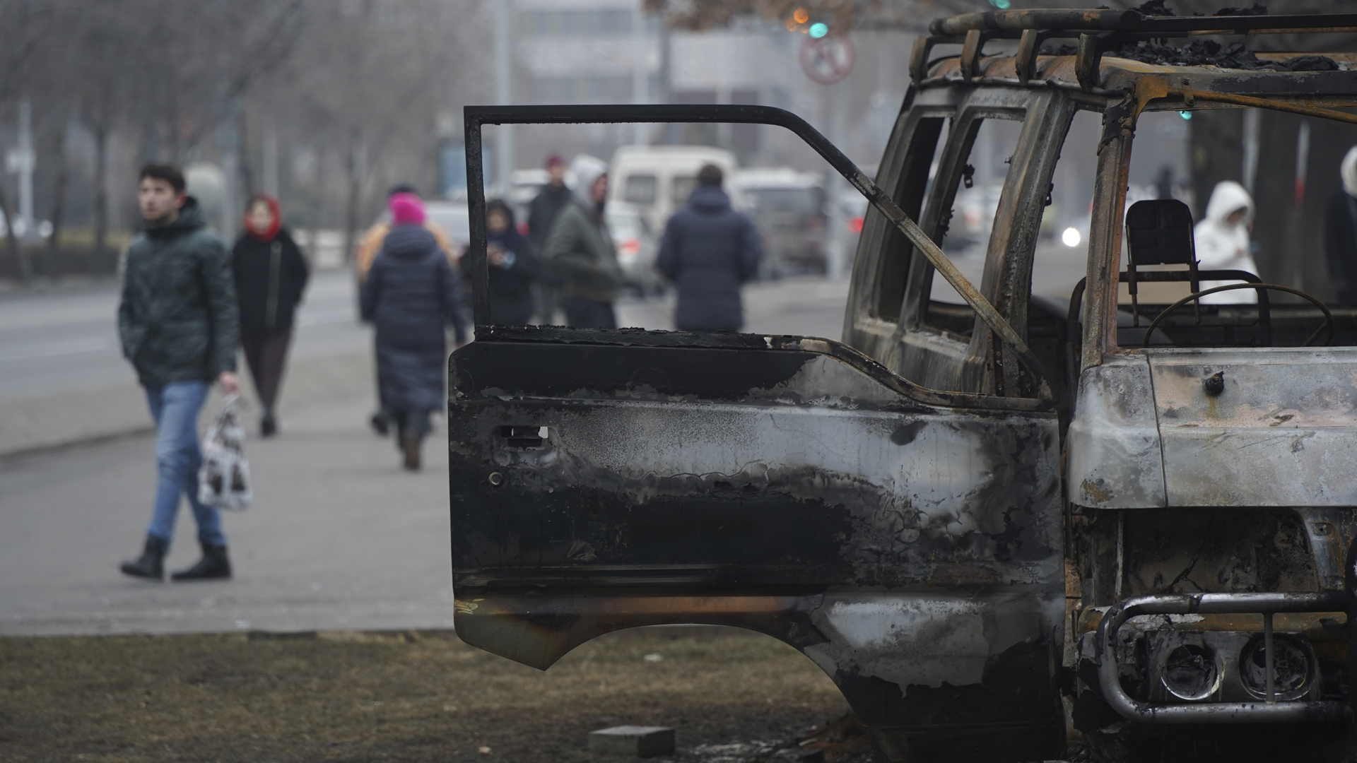 Personen gehen auf einer Straße in Almaty, auf der ein ausgebranntes Fahrzeug steht. | picture alliance/dpa/NUR.KZ/AP