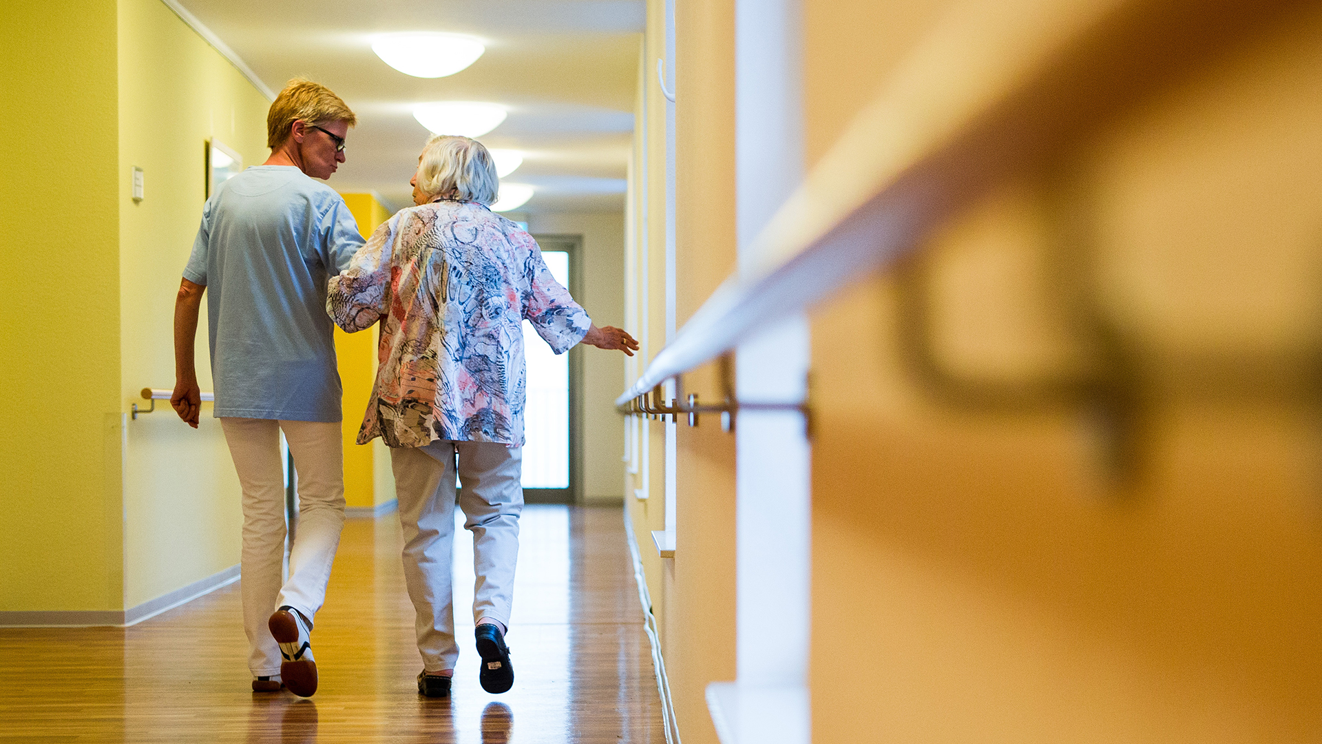Pflegerin mit einer älteren Dame gehen einen Flur entlang. (Archivbild 2018) | picture alliance/dpa
