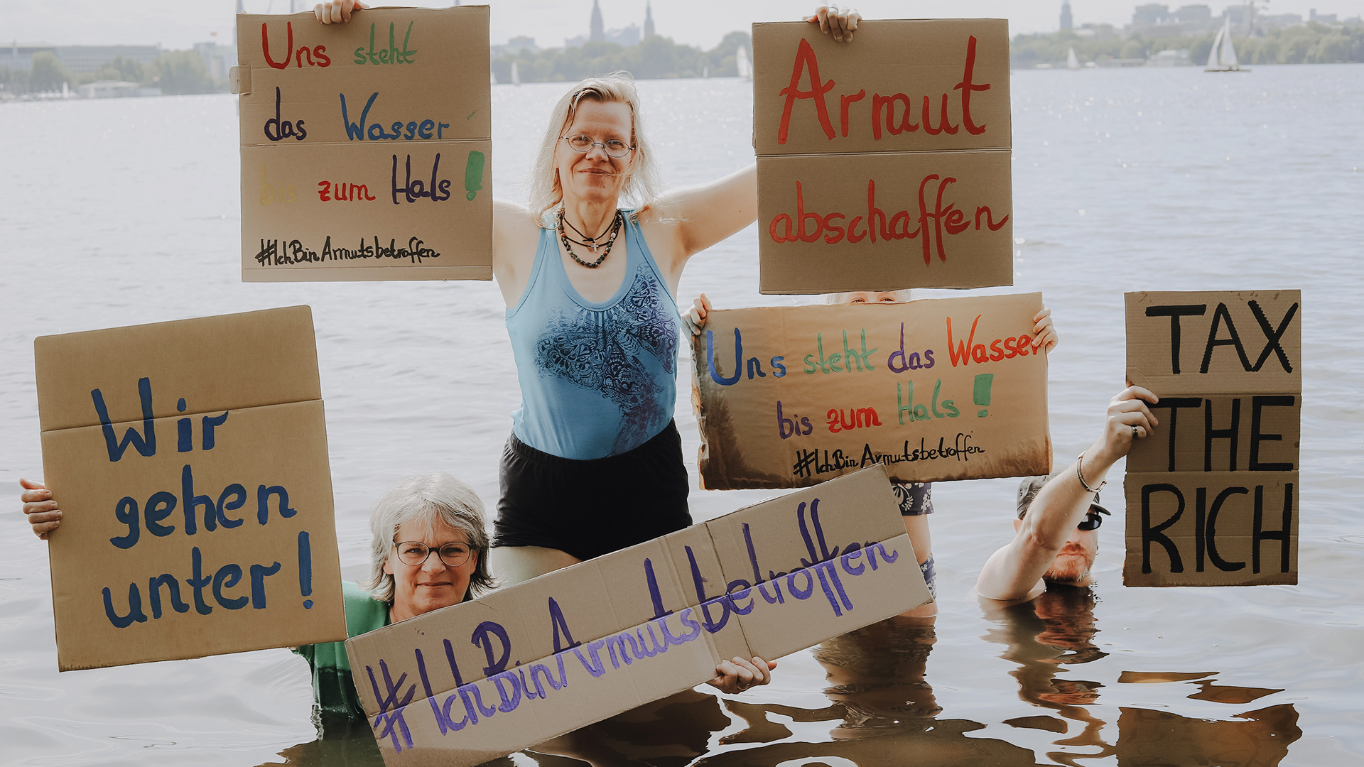 Aktivisten stehen im Wasser und halten Schilder mit der Aufschrift "#IchBinArmutsbetroffen". | OneWorryLess Foundation / Alexandra Pfeifer