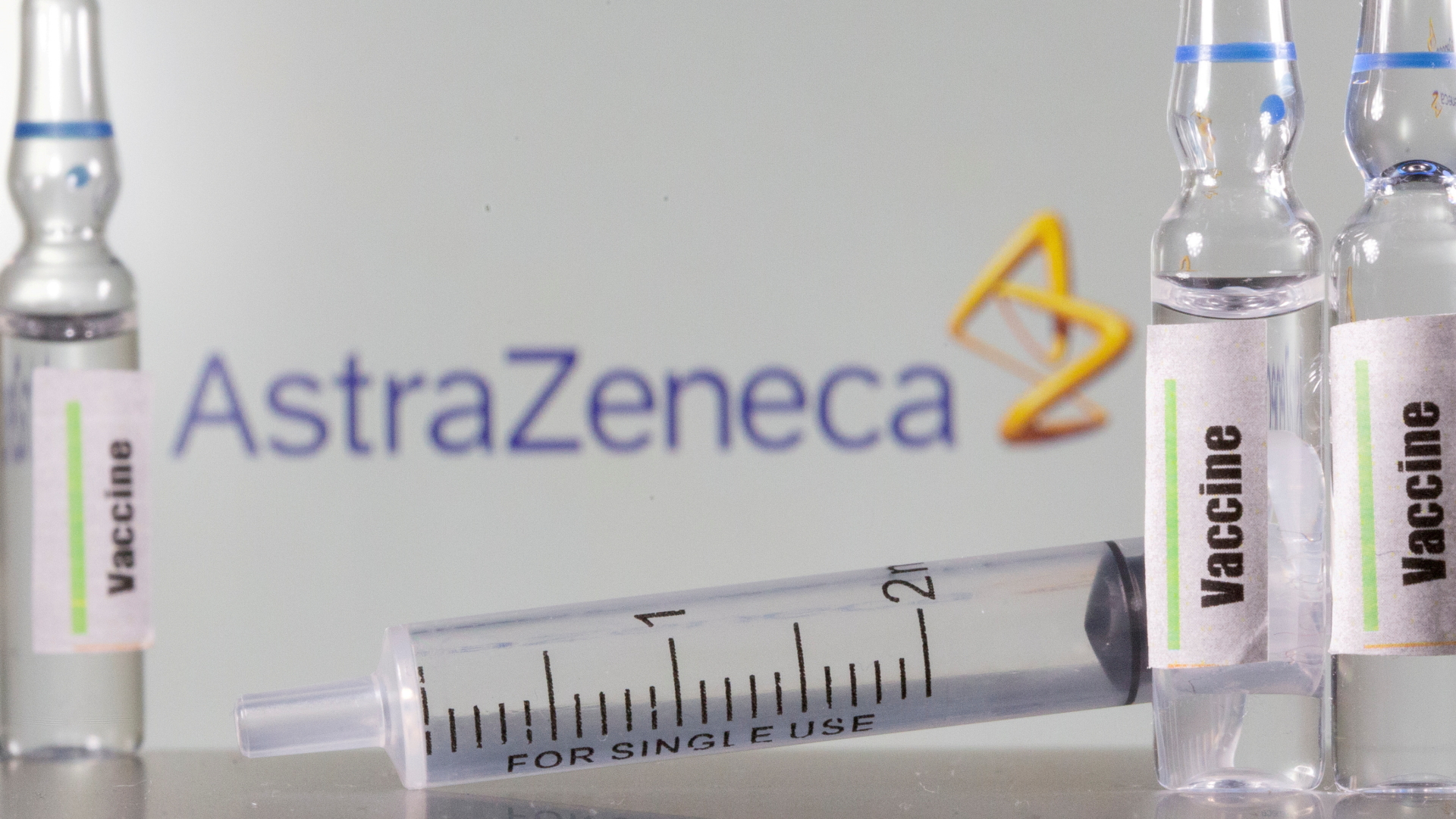 Zwischen Ampullen mit dem Corona-Impfstoff liegt eine Einmal-Spritze, im Hintergrund das Logo des Vakzin-Herstellers AstraZeneca.