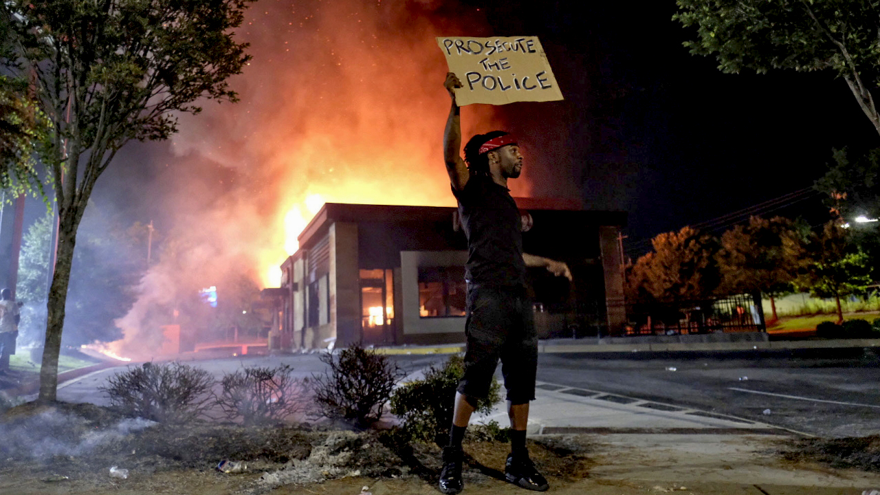 Ein Mann hält vor der brennenden Filiale einer Burgerkette in Atlanta ein Schild hoch, auf dem Strafverfolgung für die Polizei gefordert wird. | dpa