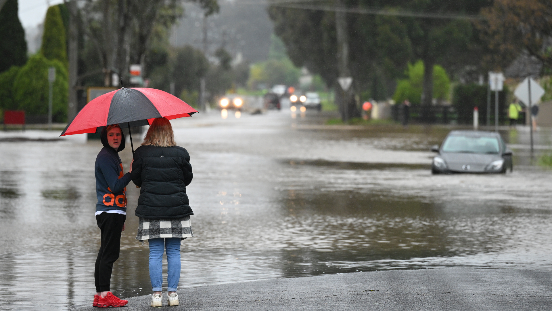 Menschen stehen an einer überschwemmten Straße und blicken auf ein im Hochwasser stehendes Auto. | dpa