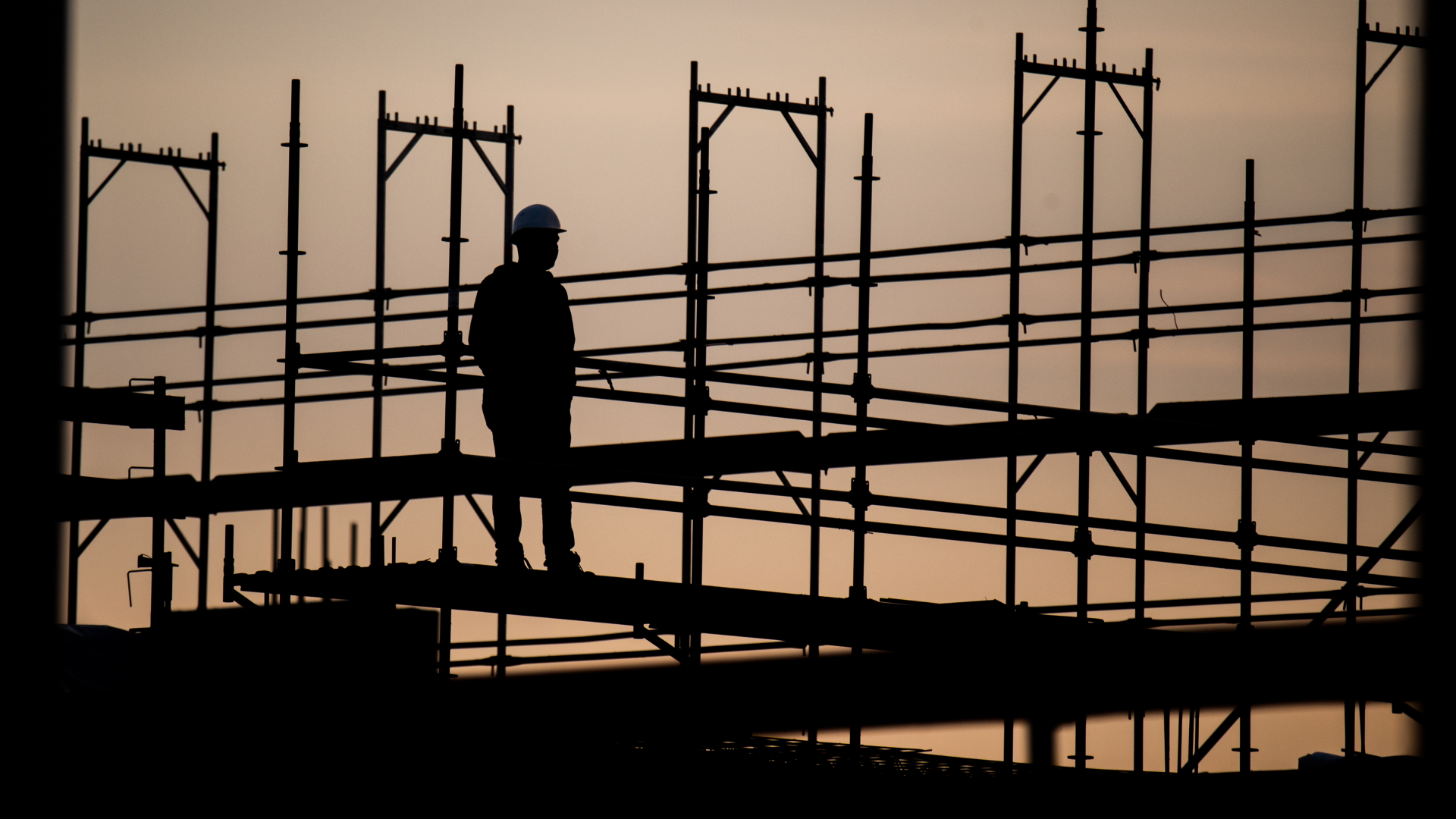 Die Silhouette eines Bauarbeiters zeichnet sich auf einer Baustelle vor dem verfärbten Morgenhimmel ab.