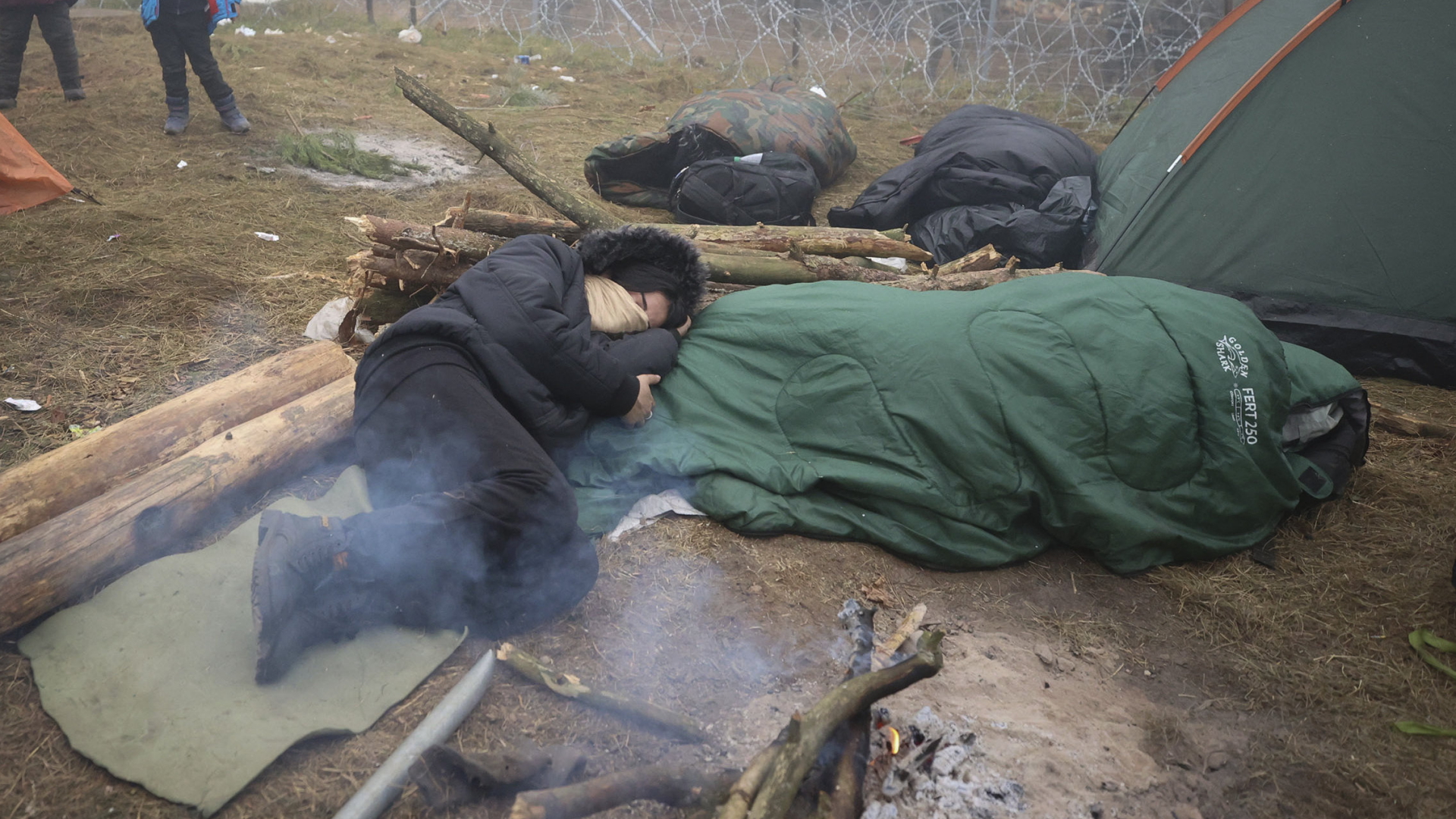 Zwei Menschen liegen in Schlafsäcken neben einem Lagerfeuer an der belarusischen Grenze zu Polen. | AP