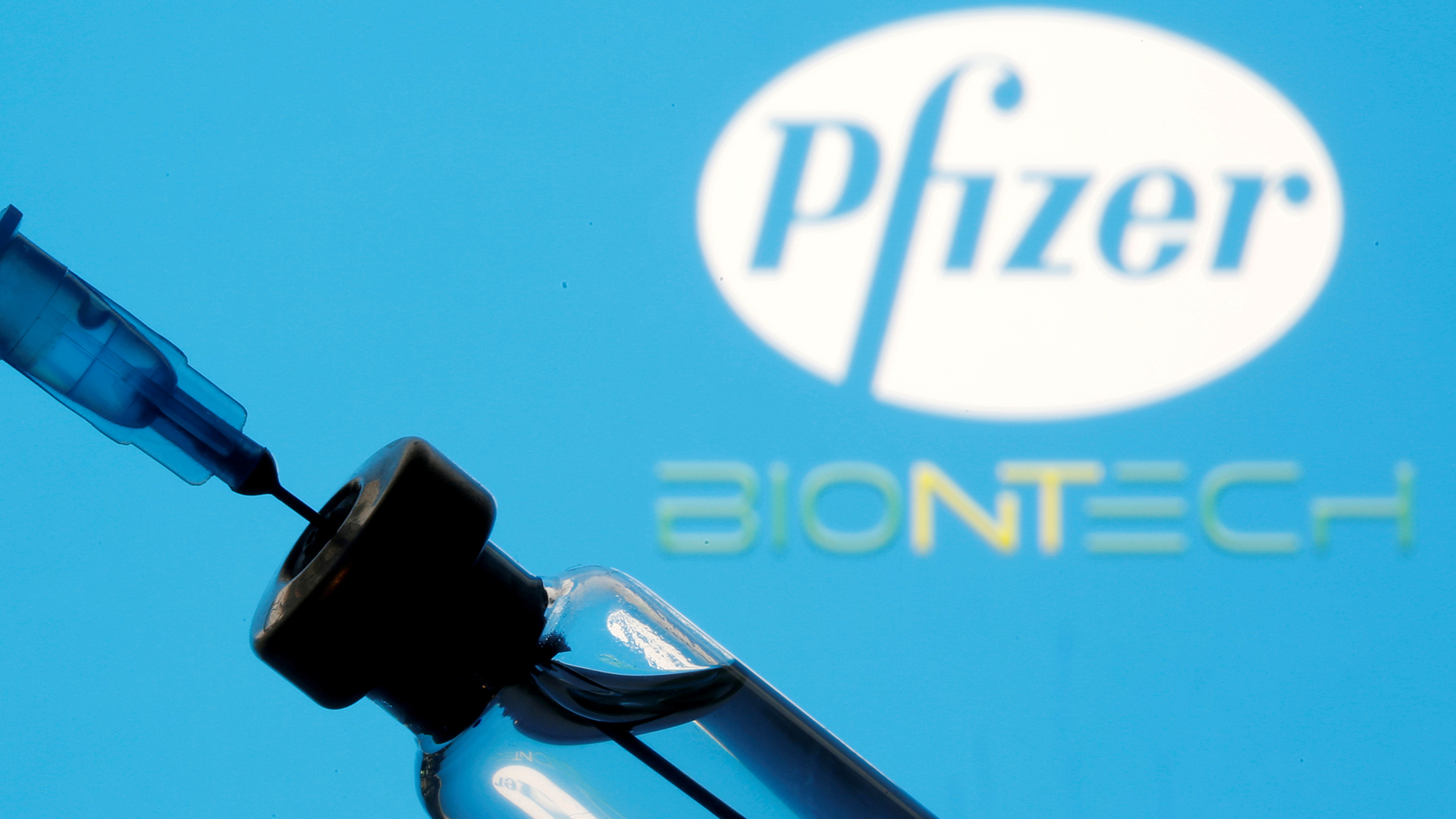Eine Spritze wird mit Impfstoff vor den Logos von Pfizer und BioNTech aufgeziogen.