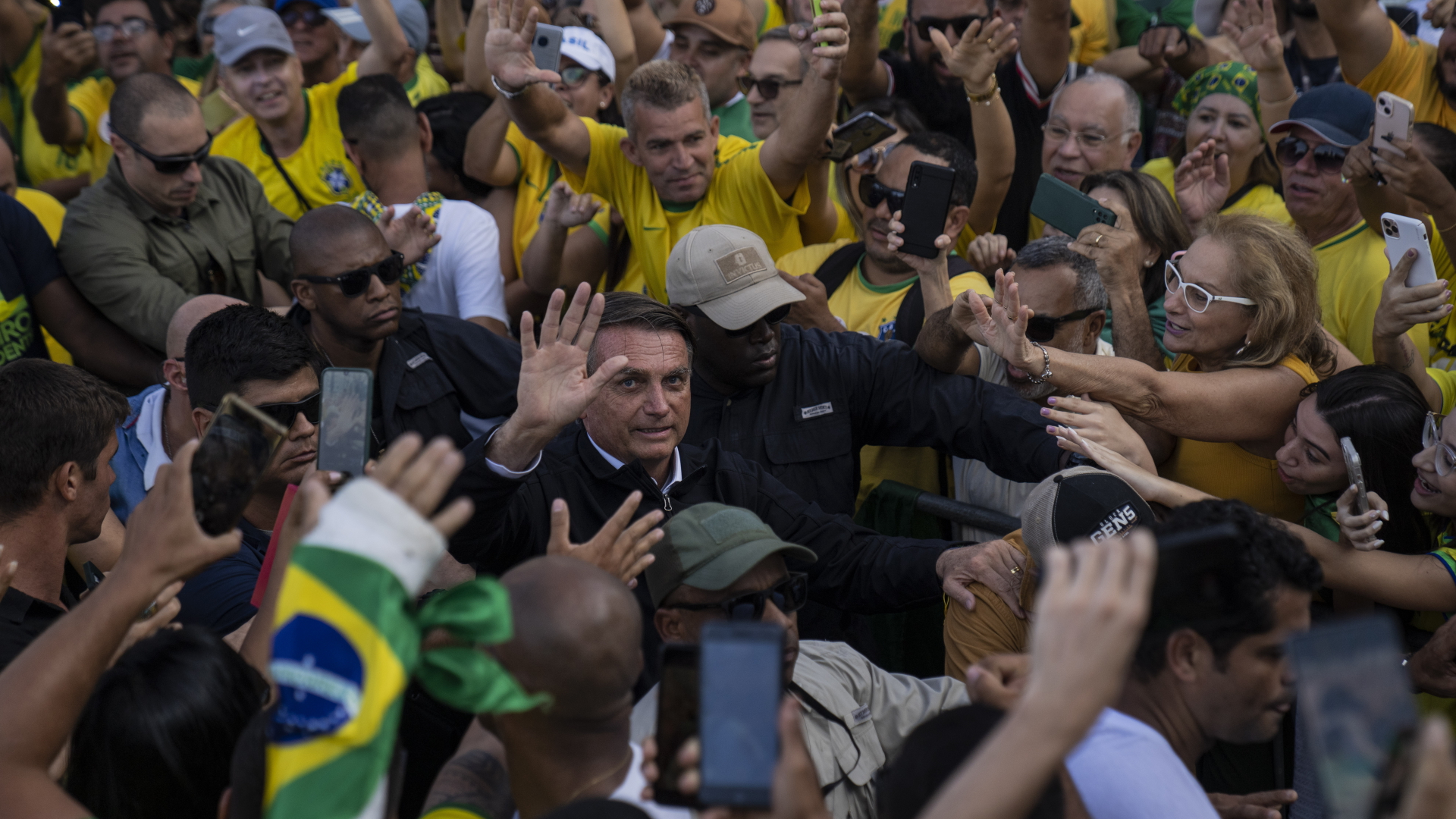 Brasiliens Präsident Jair Bolsonaro wird von einem Bodyguard durch eine Menge seiner Anhänger geführt, während der Präsident in die Kamera winkt. | dpa