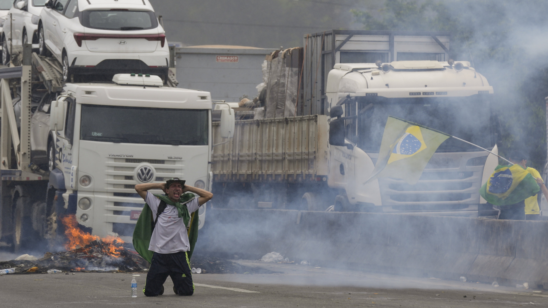 Ein Protestierender kniet mit den Händen hinter den Kopf vor einer LKW Blockade. Tränengas ist im Einsatz.  | AP