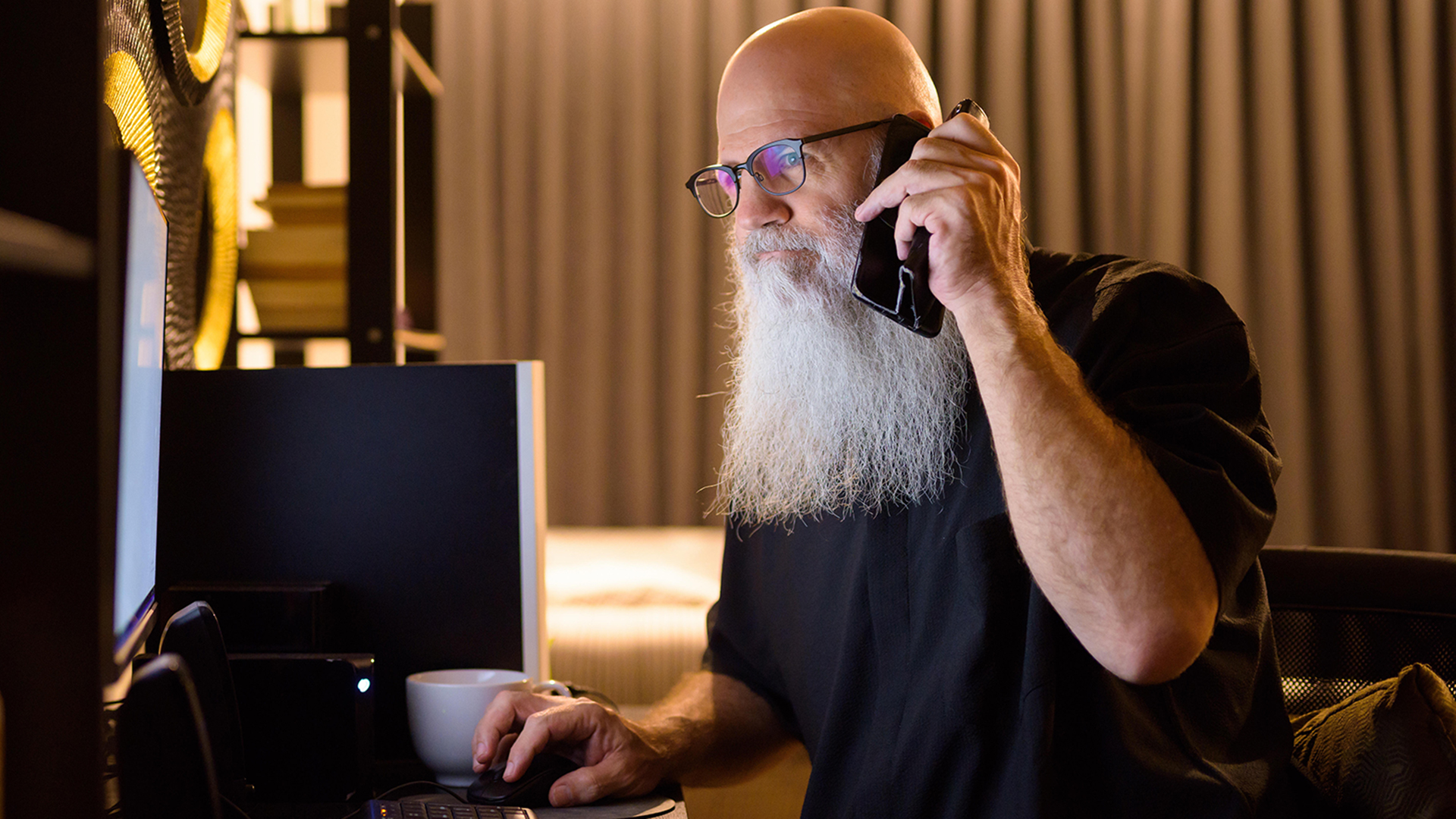 Ein Mann mit Brille und Smartphone am Ohr schaut auf einen Computer-Bildschirm | picture alliance / Zoonar