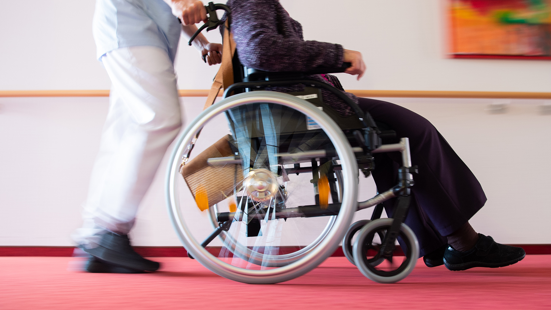 Ein Pfleger schiebt eine Bewohnerin eines Pflegeheims in einem Rollstuhl über den Flur.