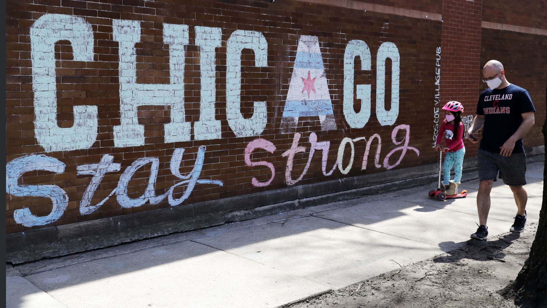 Ein Mann mit Kind, beide mit Mundschutz, gehen an einer Wand vorbei, auf die eine Künstlerin den Schriftzug "Chicago, bleib stark" gemalt hat. | AP
