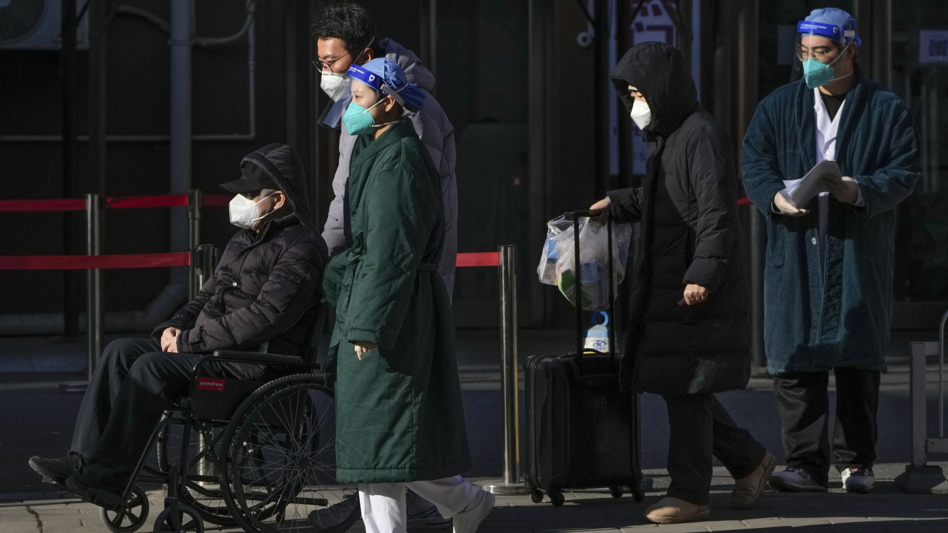 In peking schiebt ein Paar einen Mann im Rollstuhl aus einer Klinik. Alle tragen Corona-Schutzmasken. | AP