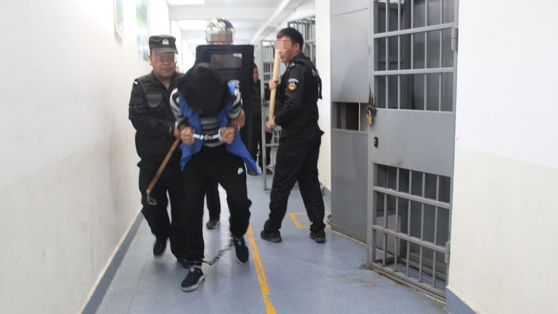 Mit Holzknüppeln bewaffnete Sicherheitskräfte führen einen Inhaftierten in Hand- und Fußfesseln ab. | Xinjiang Police Files