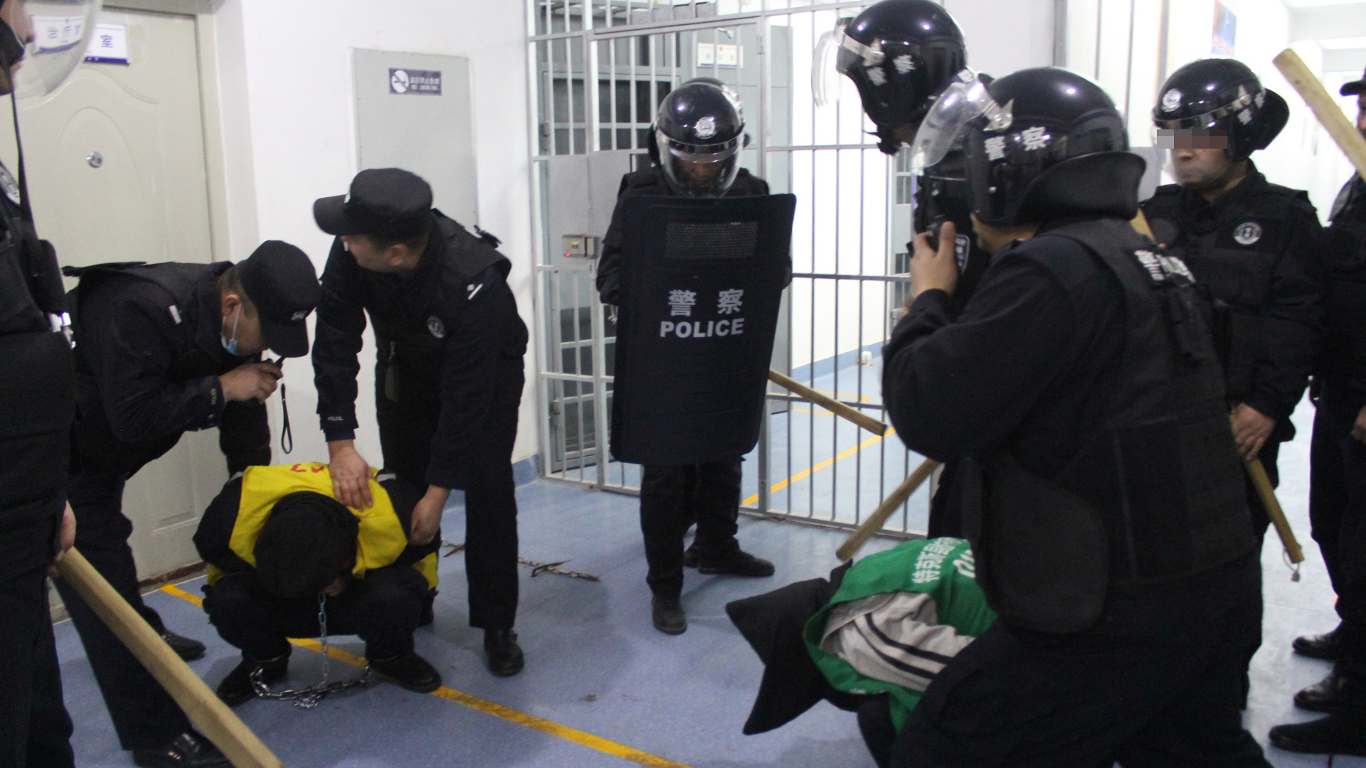 Uniformierte Menschen, bewaffnet mit Holzknüppeln, legen einem Inhaftierten Handschellen an. | Xinjiang Police Files