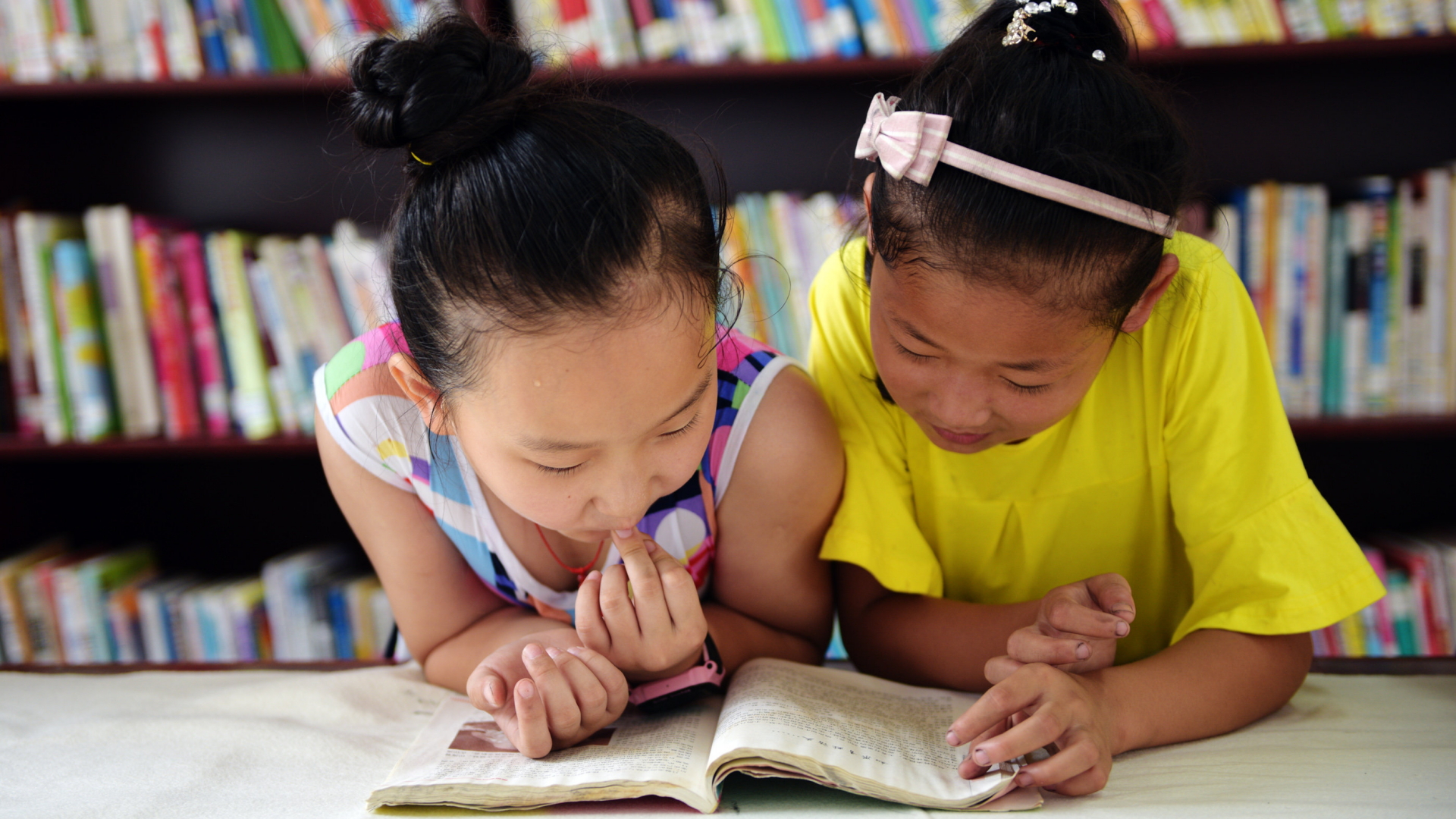 Zwei chinesische Mädchen beugen sich in einer Bibliothek über ein aufgeschlagenes Buch.