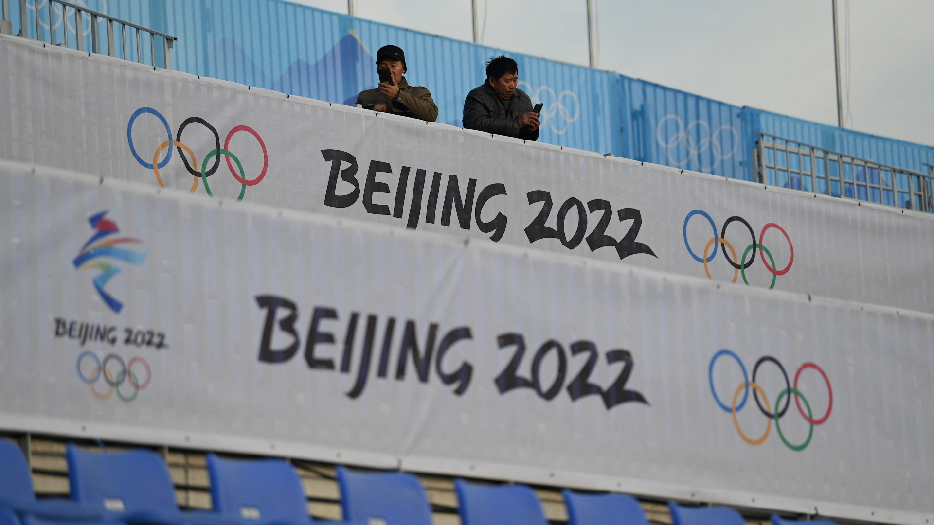 Zwei Männer mit Handys stehen hinter einem Plakat der Olympischen Spiele | AFP