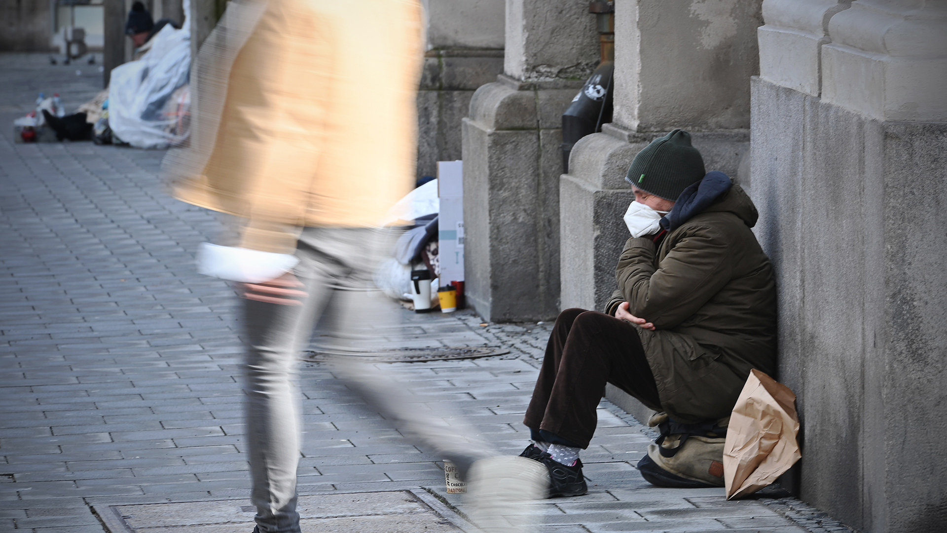 Ein Bettler sitzt mit Mundschutz auf dem Gehweg.
