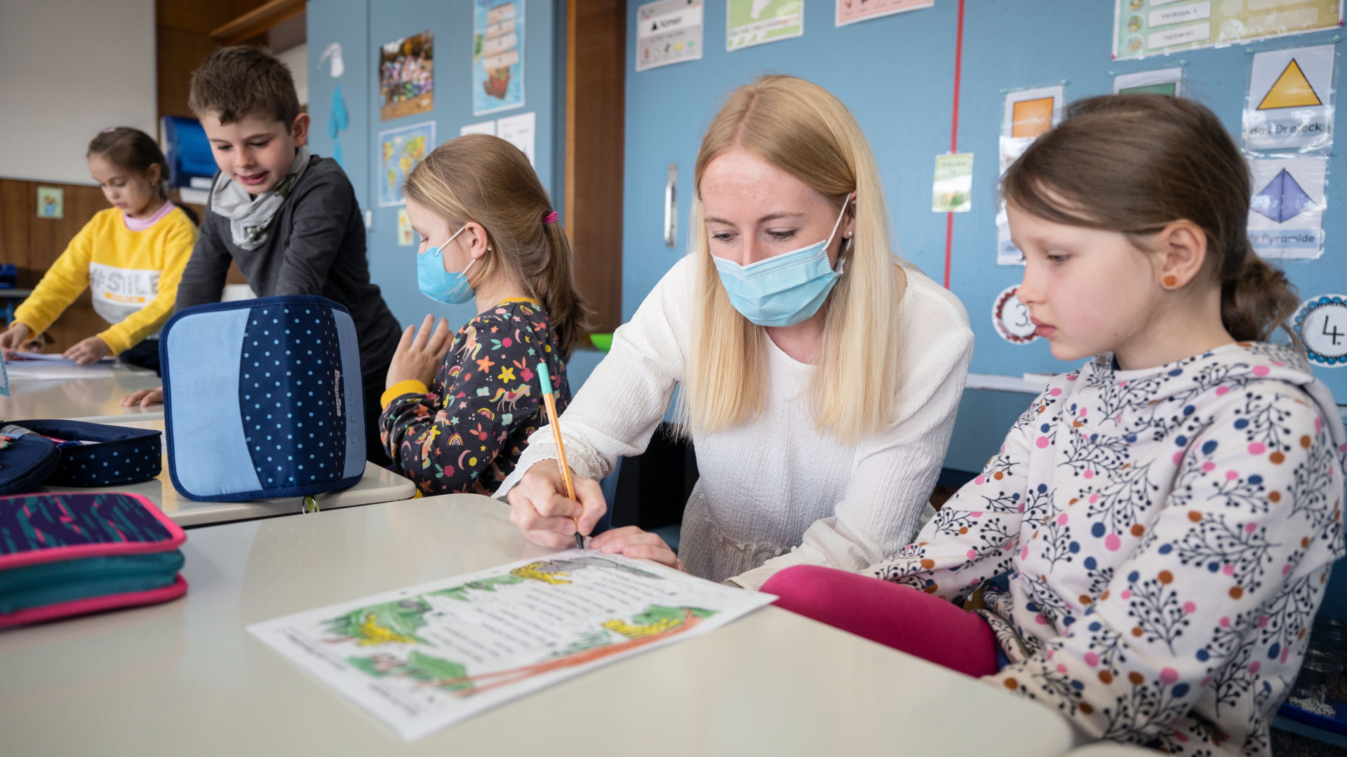 Eine Lehrerin mit Mund-Nasen-Schutz sitzt neben Schulkindern am Schreibtisch.