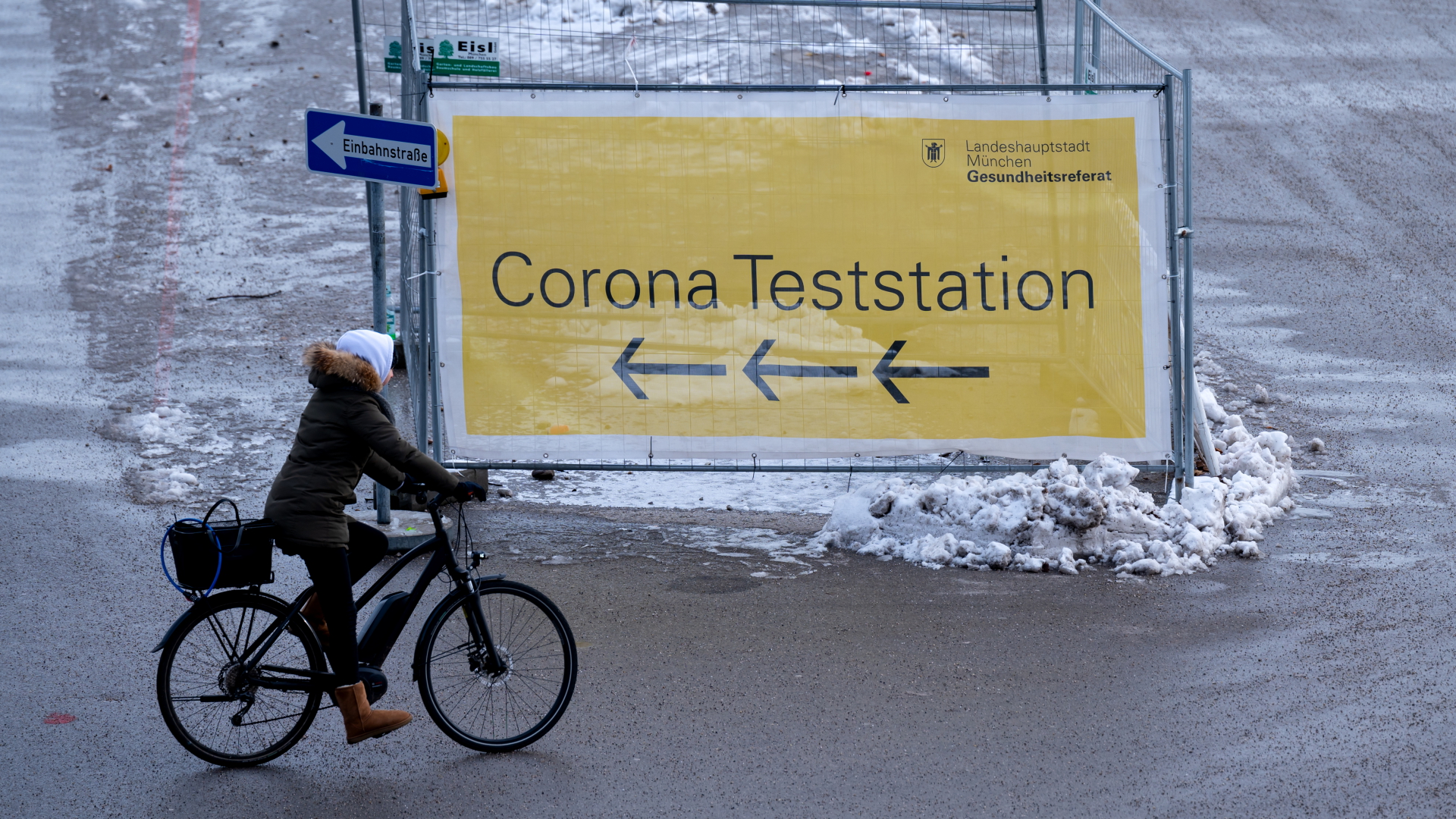Eine Frau fährt mit ihrem Fahrrad auf der Theresienwiese an einem Plakat mit der Aufschrift "Corona Teststation" entlang.