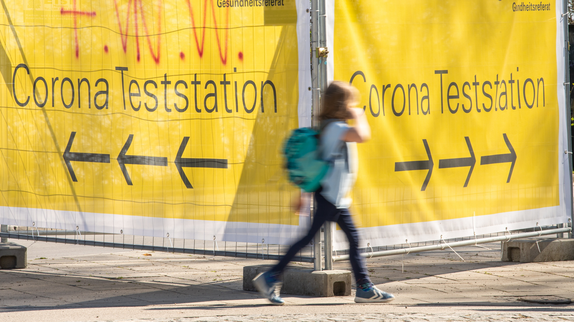 Ein Junge läuft an zwei großen, gelben Bannern vorbei, die den Weg zu einer Corona-Teststation weisen. | dpa