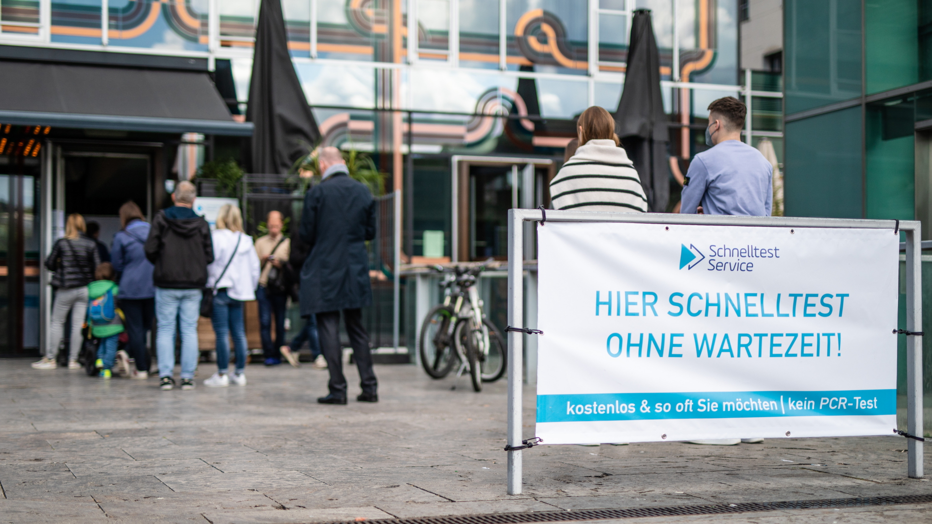 Menschen stehen vor einem Stuttgarter Testzentrum neben einem Transparent mit der Aufschrift "Hier Schnelltest - Ohne Wartezeit" an. | dpa