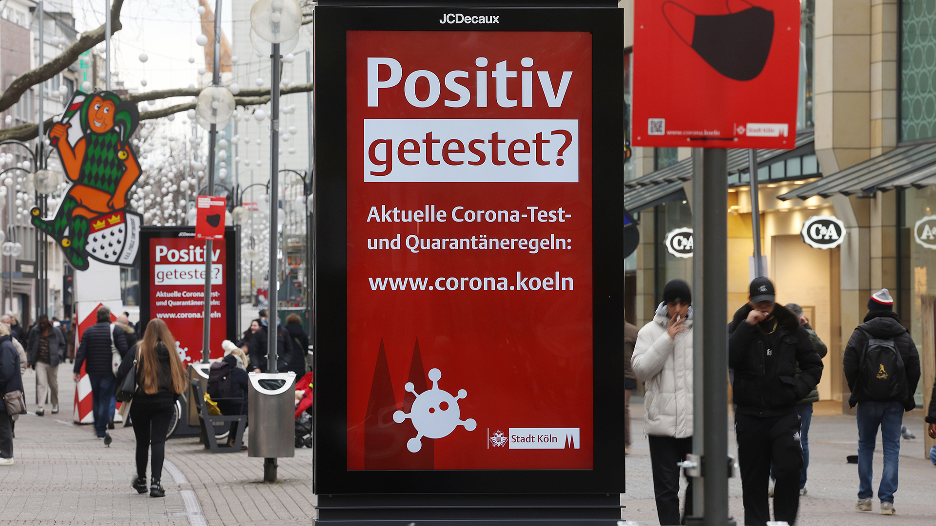 Menschen gehen an einer elektronischen Werbetafel mit der Aufschrift "Positiv getestet?" in der Kölner Innenstadt vorbei.  | dpa