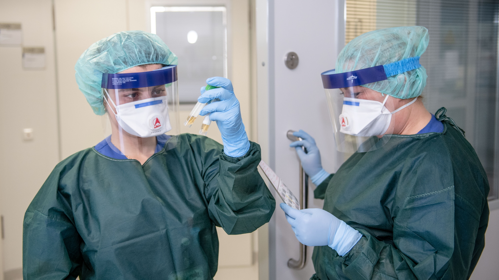 Krankenschwestern auf der Infektionsstation der Uniklinik Essen betrachten in Schutzkleidung und mit einer Atemmaske zwei Abstrichröhrchen.