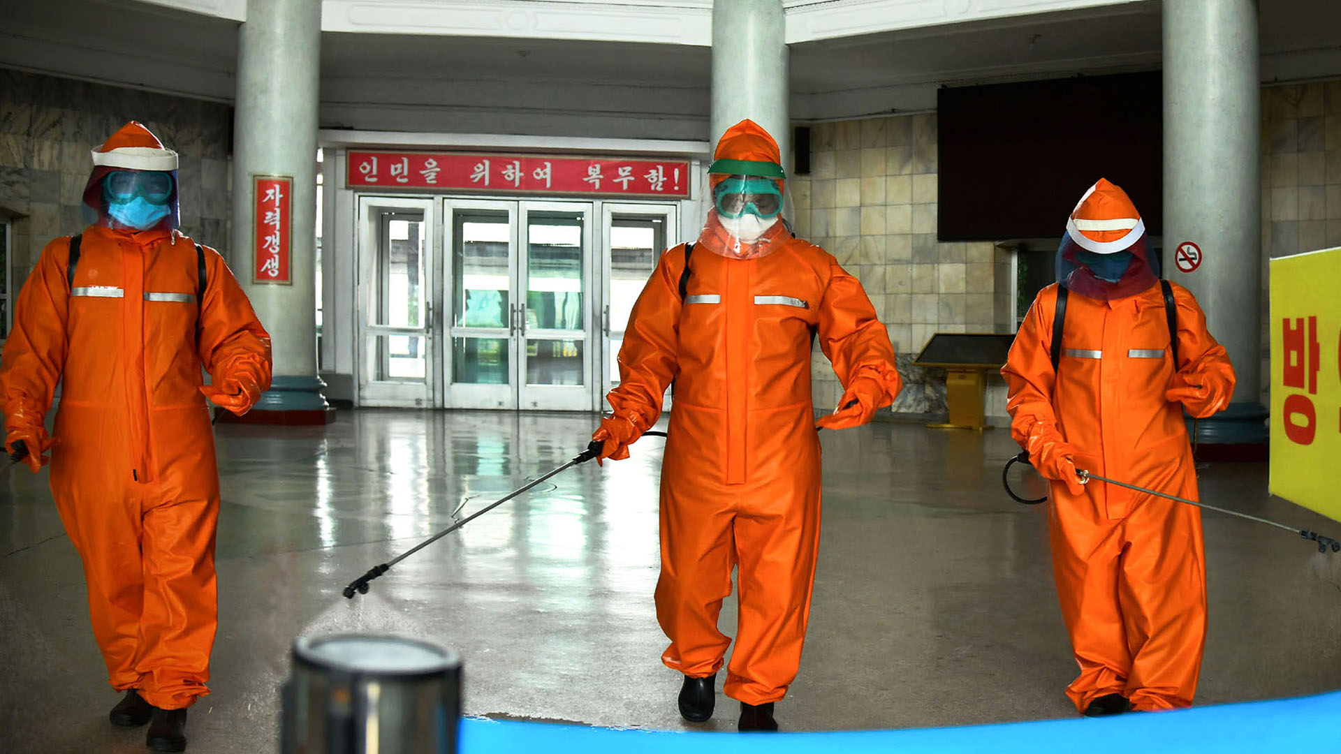 Drei Personen in orangefarbenen Schutzanzügen desinfizieren ein Bahnhofsgebäude. | picture alliance/dpa/kcna