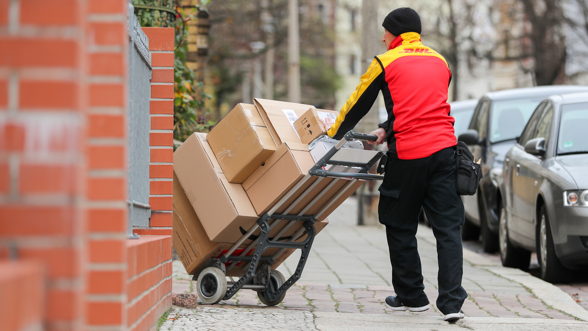 Ein Paketzusteller von DHL geht mit einer Sackkarre voller Pakete zu einem Haus. | dpa