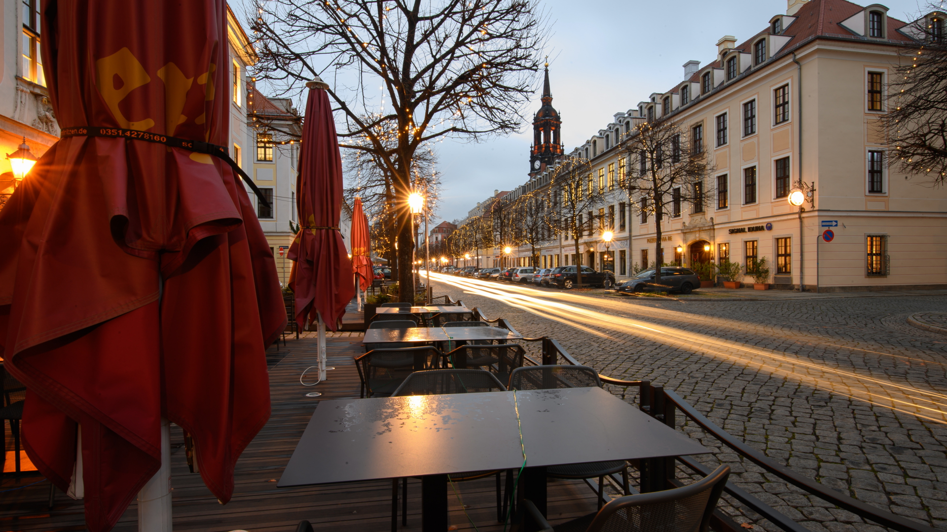 Leere Tische und geschlossene Schirme stehen vor einem Restaurant in Dresden, während auf der Königstraße die Scheinwerfer vorbeifahrender Autos Lichtspuren ziehen. | dpa