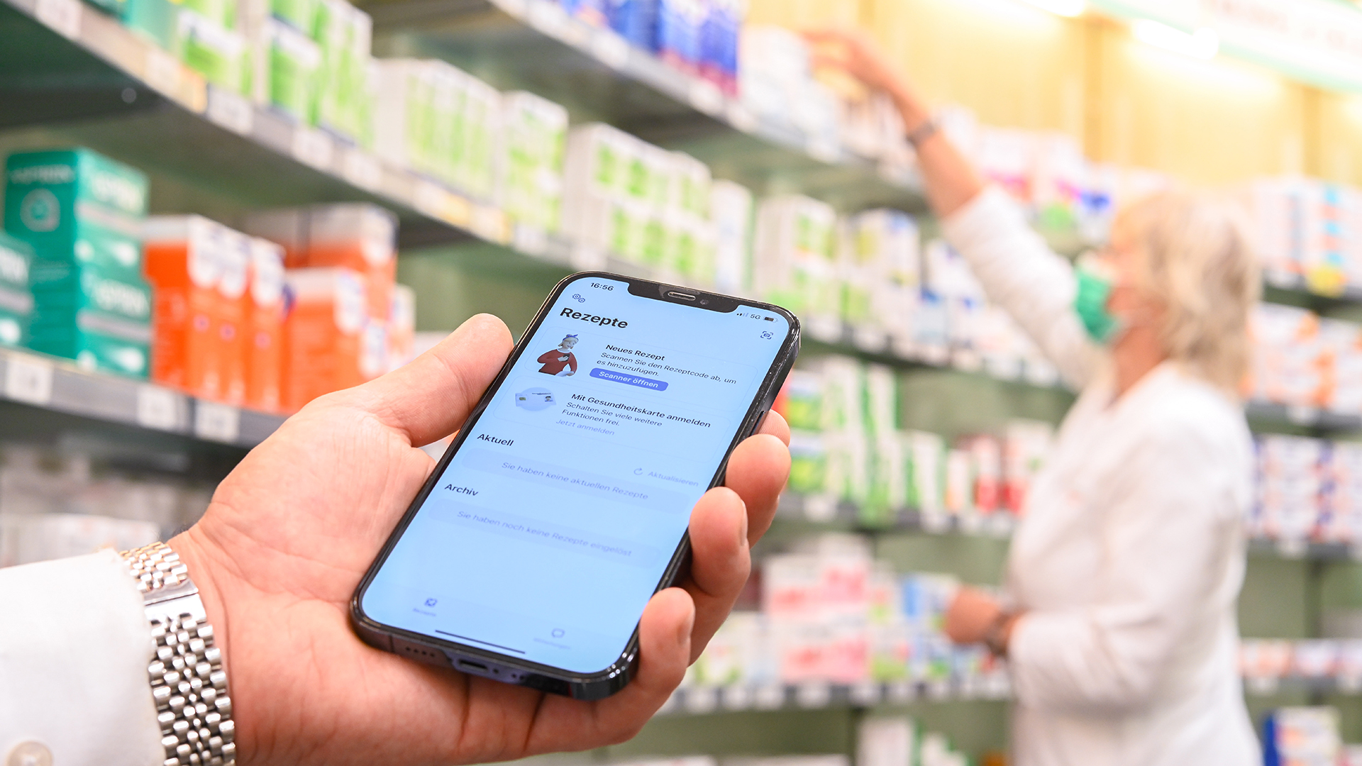 Auf einem Smartphone ist die geöffnete App "Das E-Rezept" zu sehen, während im Hintergrund eine Apothekerin in einer Apotheke an einem Regal steht.  | dpa