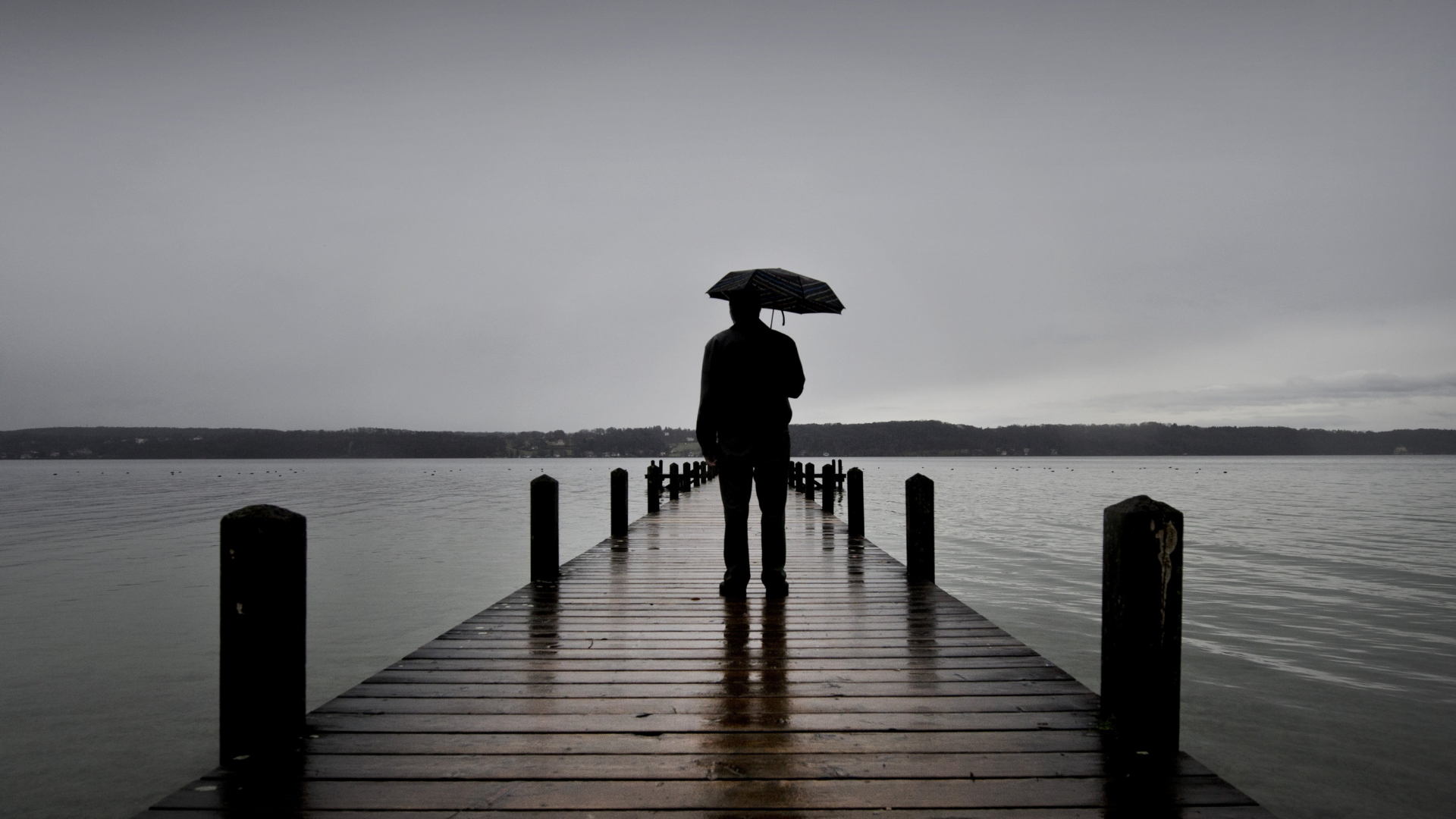 Auf einem regennassen Bootssteg ist vor grauem Himmel ein Mann mit Regenschirm zu sehen.