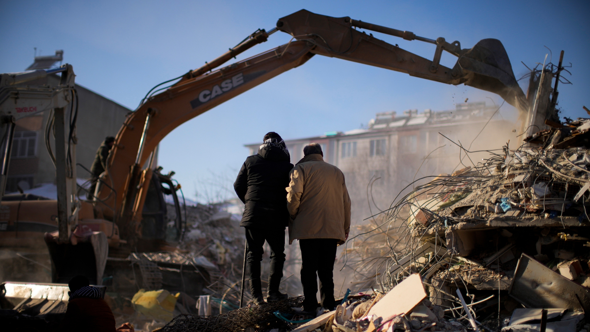 Zwei Personen schauen dabei zu, wie Rettungsteams in einem zerstörten Gebäude nach Menschen suchen. | dpa