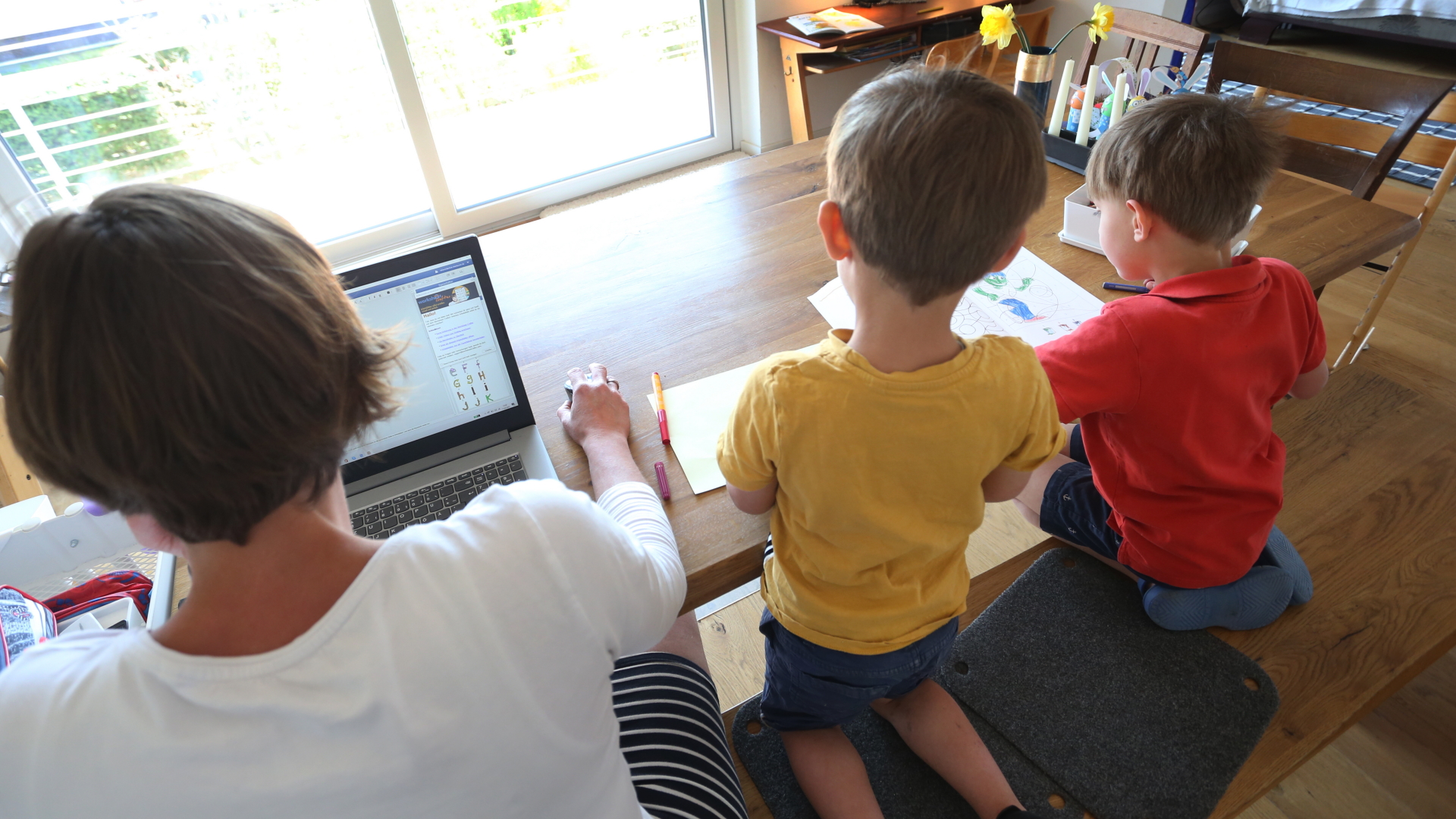 Eine Mutter sitzt am Laptop, neben ihr zeichnen ihre beiden kleinen Söhne.