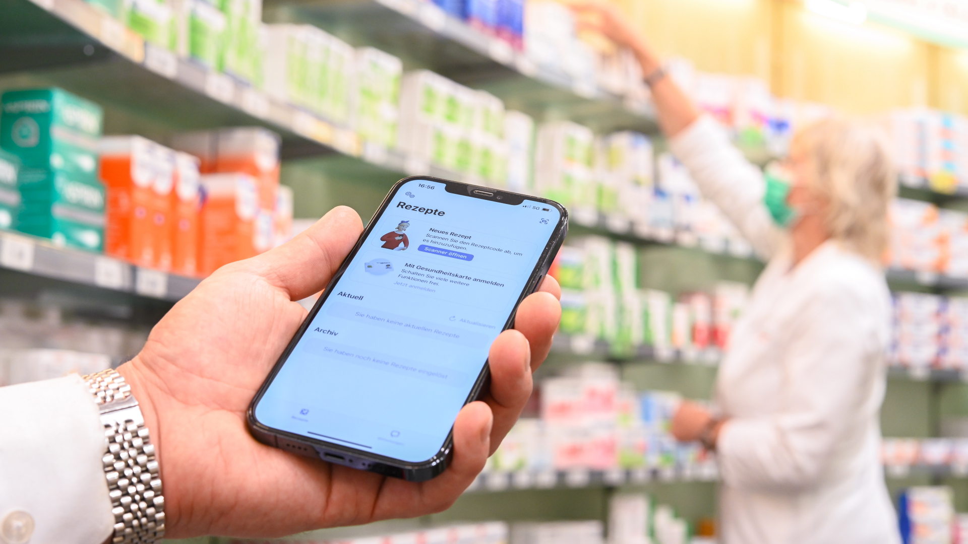 Auf einem Handy ist die E-Rezept-App geöffnet, im Hintergrund sucht eine Apothekerin in einem Regal ein Medikament. | dpa