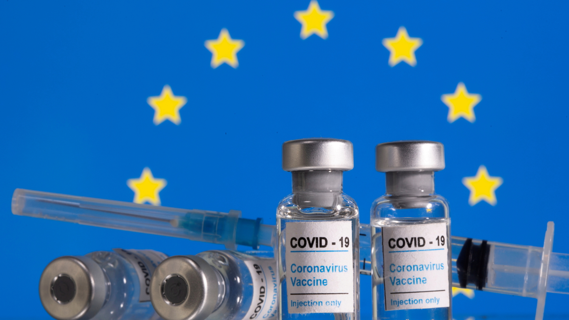 Mehrere Impullen mit einem Corona-Impfstoff und eine Spritze vor einer Europa-Flagge. | REUTERS