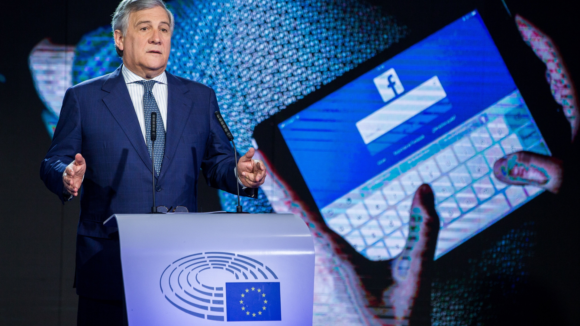 EU-Parlamentspräsident Tajani hält eine Rede, im Hintergrund zeigt ein Foto Hände die sich auf einem Tablet bei Facebook einloggen.