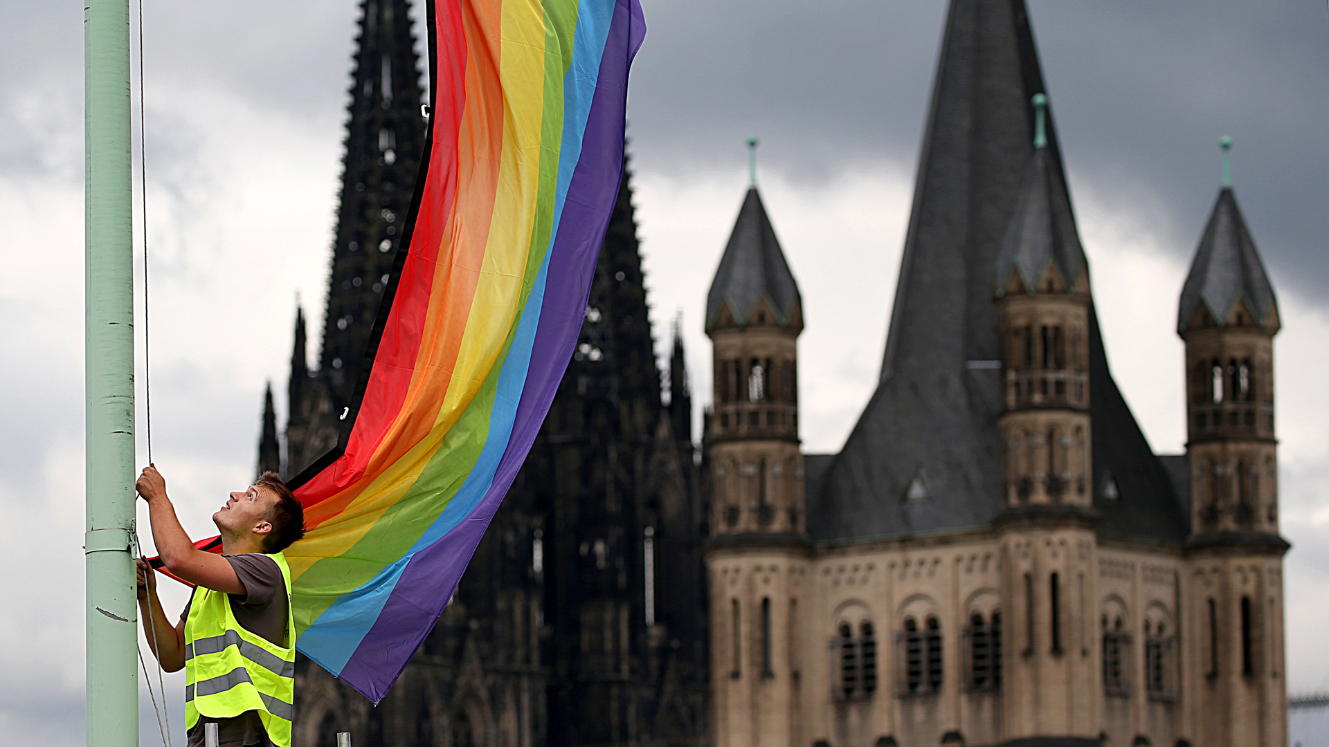 Ein Arbeiter hängt vor der Kulisse des Doms und der Kirche Groß St. Martin eine Regenbogenfahne auf. | picture alliance/dpa