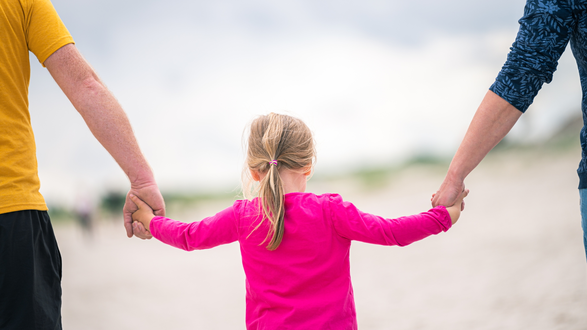 Ein Mädchen läuft Hand in Hand mit ihren Eltern am Strand entlang. | dpa