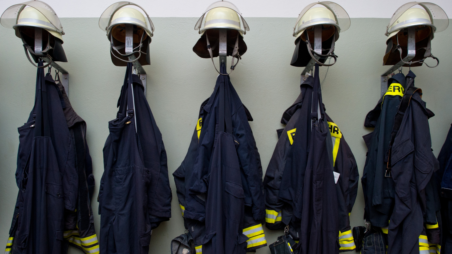 Feuerwehruniformen und -helme hängen aufgereiht an einer grauen Wand. | dpa