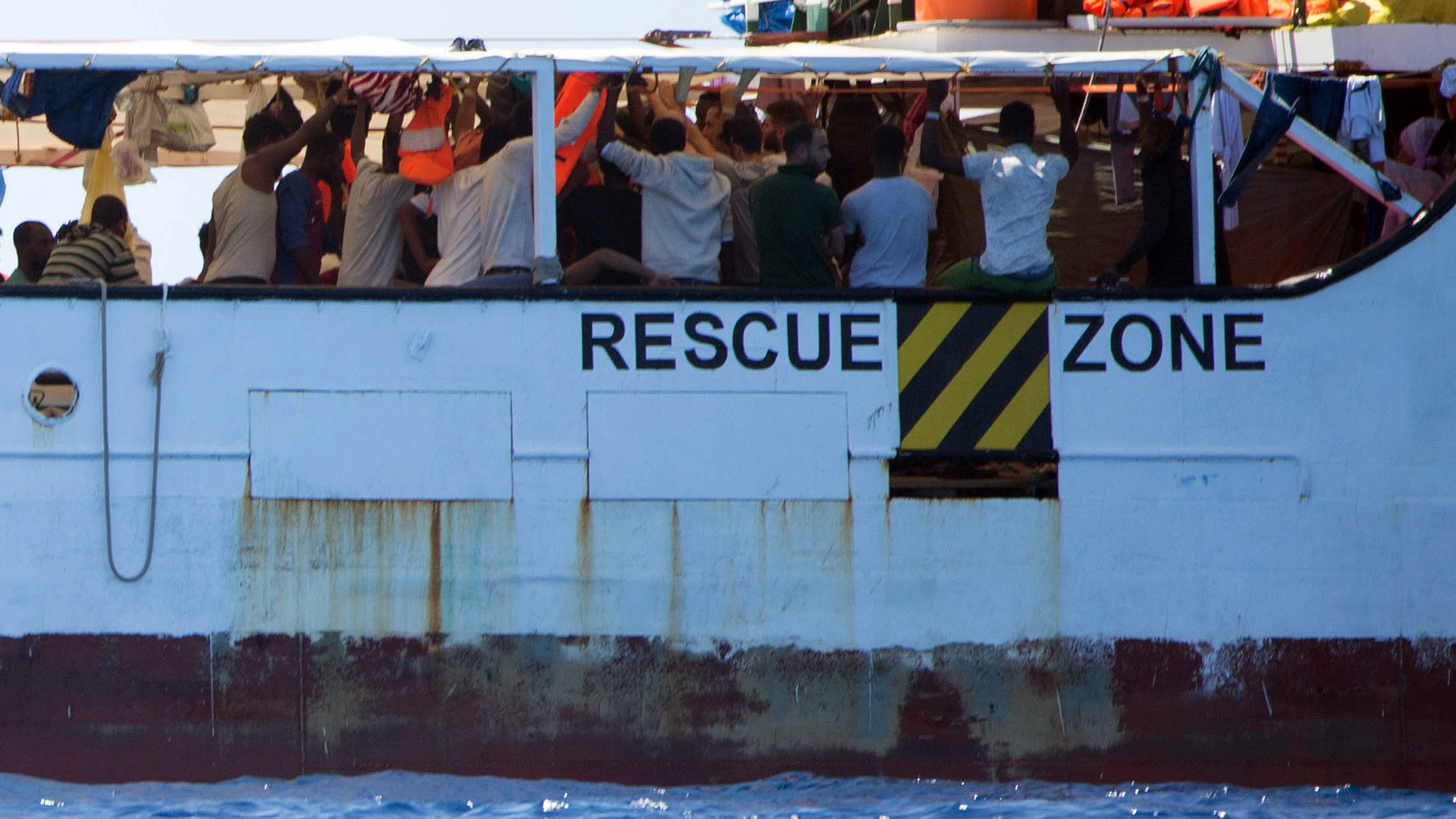 Aus dem Meer gerettete Migranten stehen auf dem Deck des Rettungsschiffes "Open Arms" der Hilfsorganisation Proactiva Open Arms, an der Bordwand steht der Schriftzug "Rescue Zone" (Rettungszone) (Archivbild). | dpa