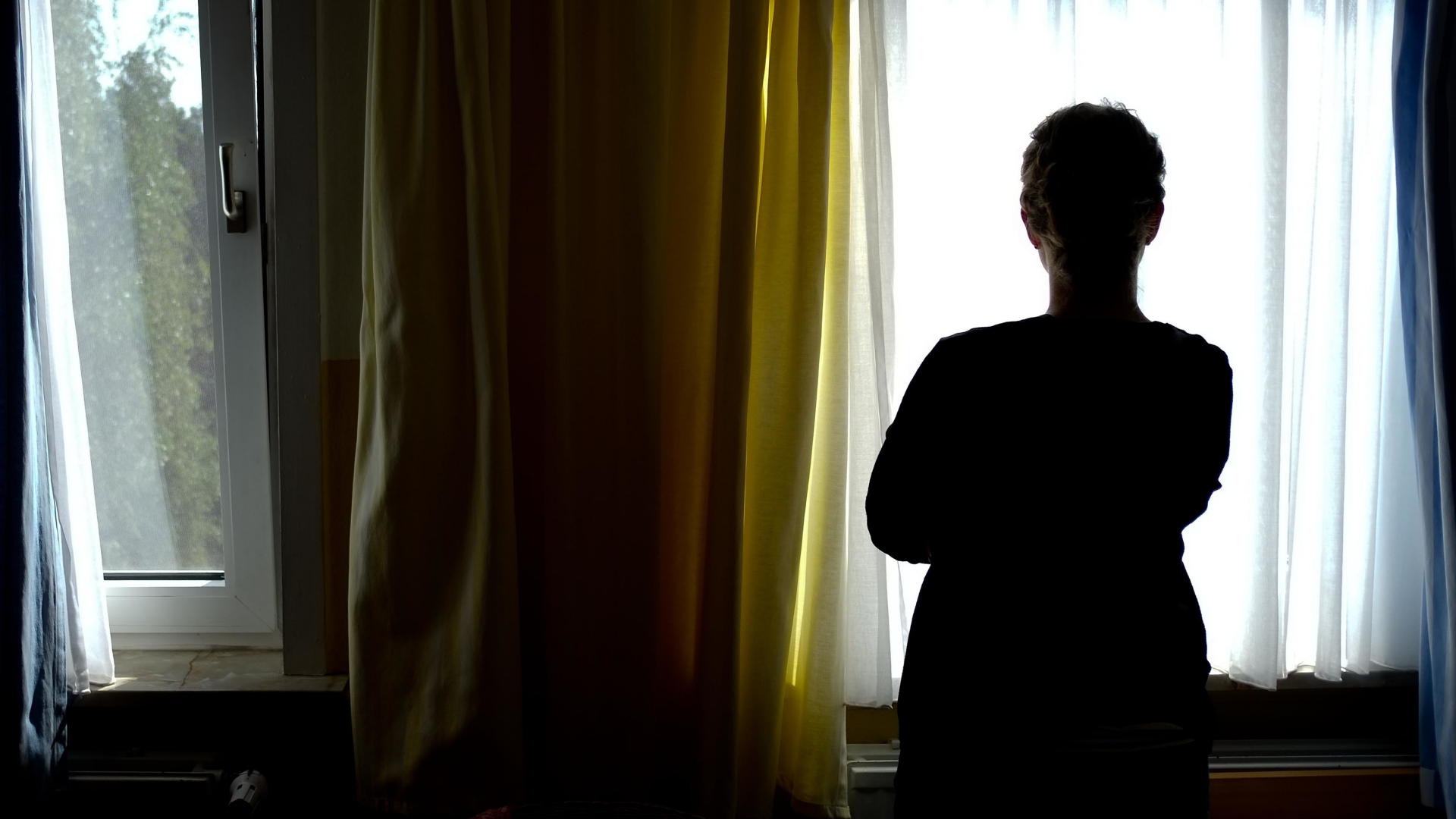 Eine Frau steht vor einem Fenster und ist im Gegenlicht nur als dunkle Silhouette erkennbar.