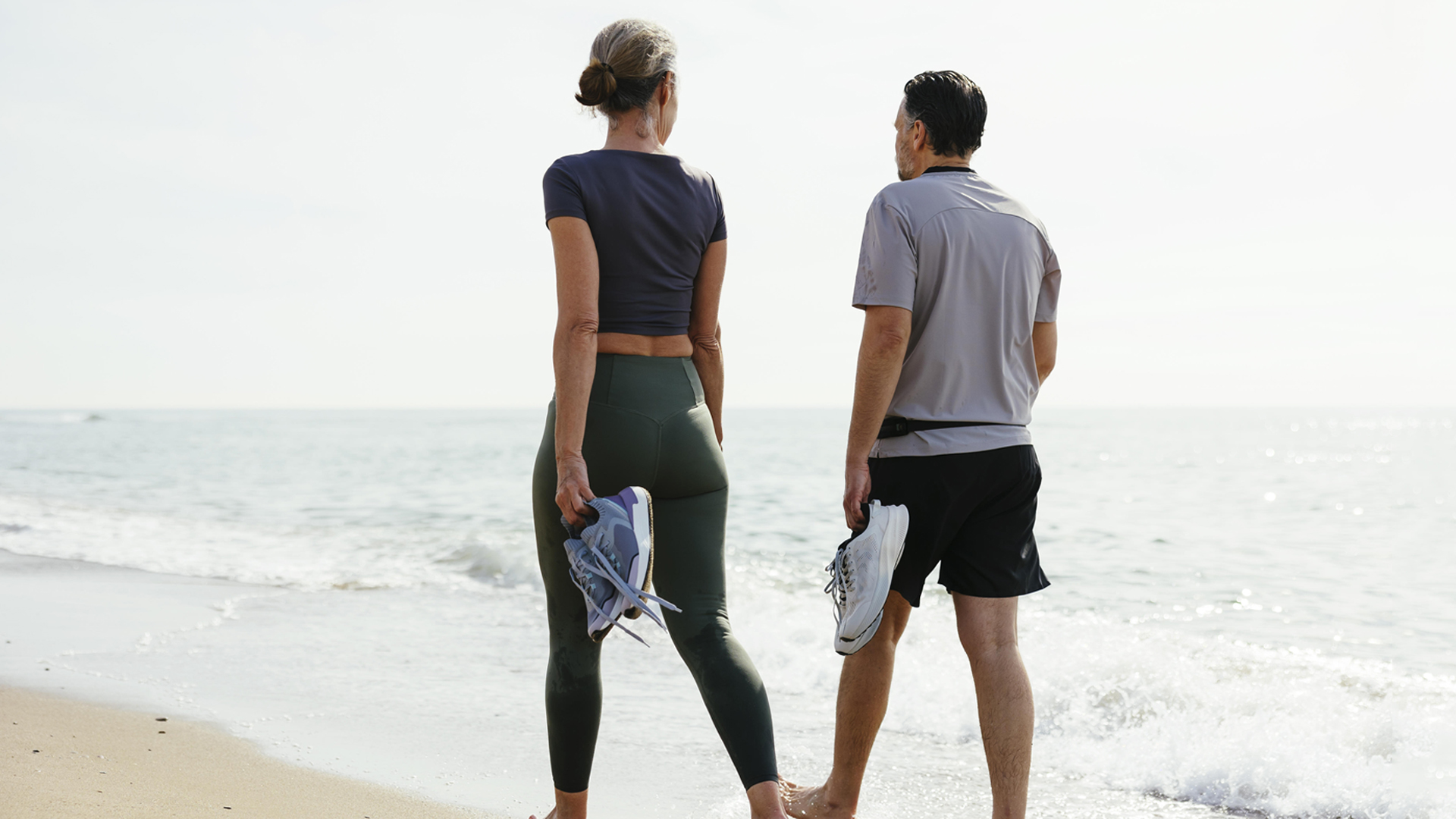 Eine Frau und eine Mann gehen barfuß am Strand entlang - in den Händen halten sie Sportschuhe - Rückenansicht