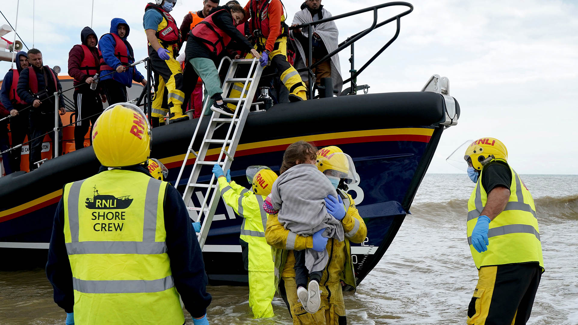 Ein junges Mädchen wird ans Ufer getragen, während eine Gruppe von Menschen, bei denen es sich vermutlich um Migranten handelt, nach Dungeness gebracht wird. | dpa