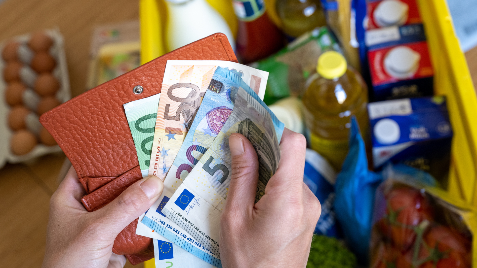 Hände ziehen einige Geldscheine aus einem Portemonnaie, darunter verschwommen ein gefüllter Einkaufskorb.